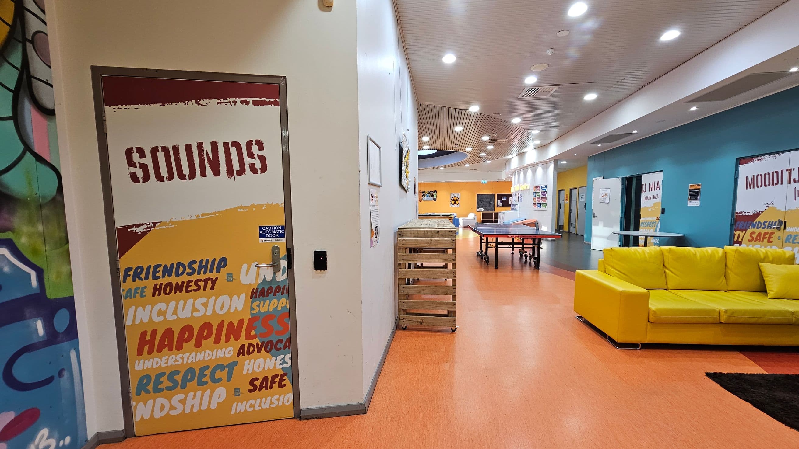 A brightly lit indoor space with an orange floor and a white ceiling featuring recessed lights. On the left, a door labelled "SOUNDS" is decorated with words like "FRIENDSHIP" and "HAPPINESS." Adjacent is a wall with colourful graffiti. In the centre, a wooden crate and a ping pong table are visible. On the right, a vibrant yellow sofa sits against a blue wall with more labelled doors. The corridor extends into the distance, with various posters and signs on the walls.