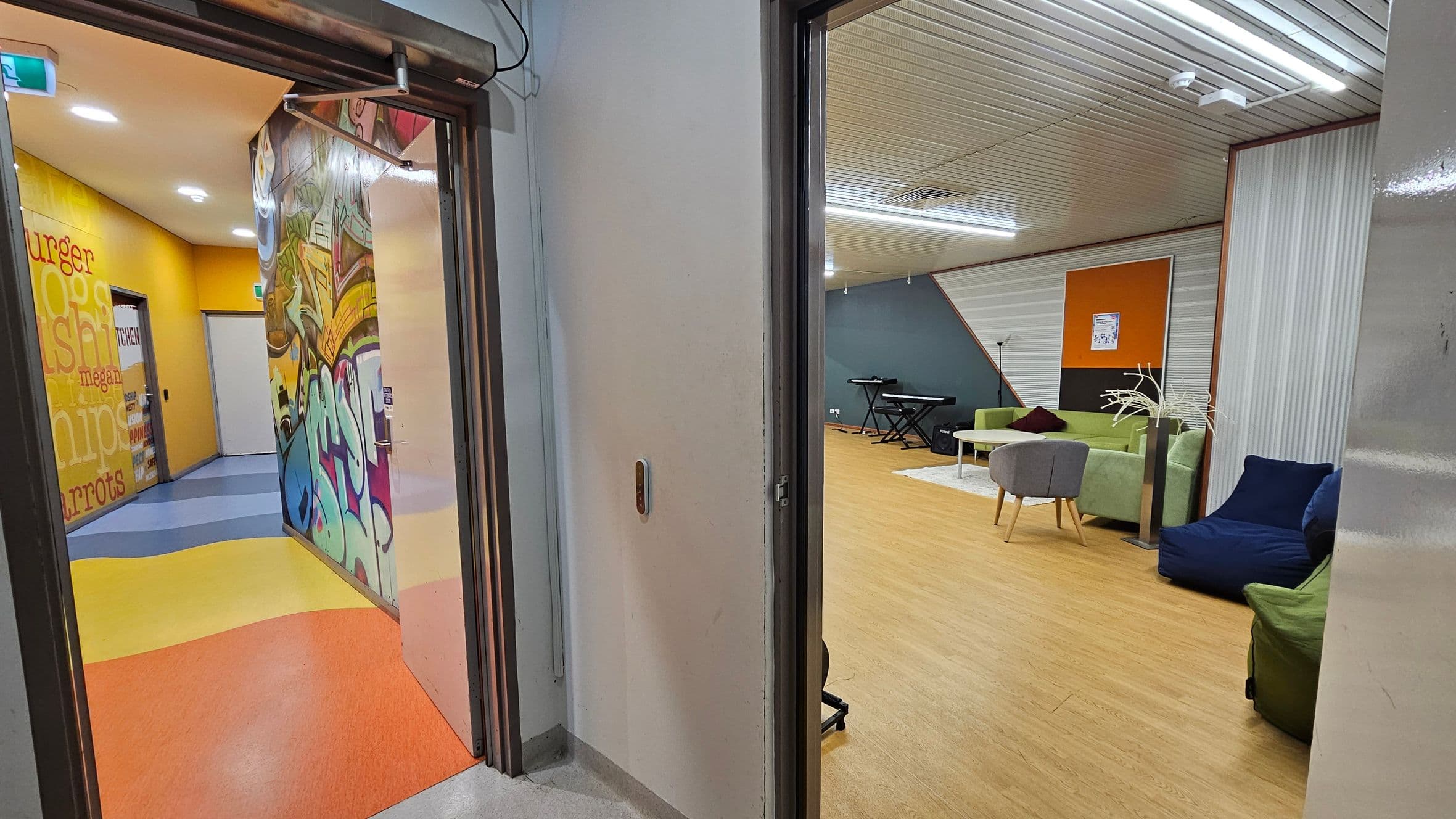 On the left, a hallway with vibrant graffiti art and a yellow wall featuring words like "burger" and "chips." The floor is a mix of blue, yellow, and orange. On the right, a room with a wooden floor, green and blue seating, a grey chair, and a keyboard against a blue wall. The ceiling is lined with white panels and fluorescent lights.