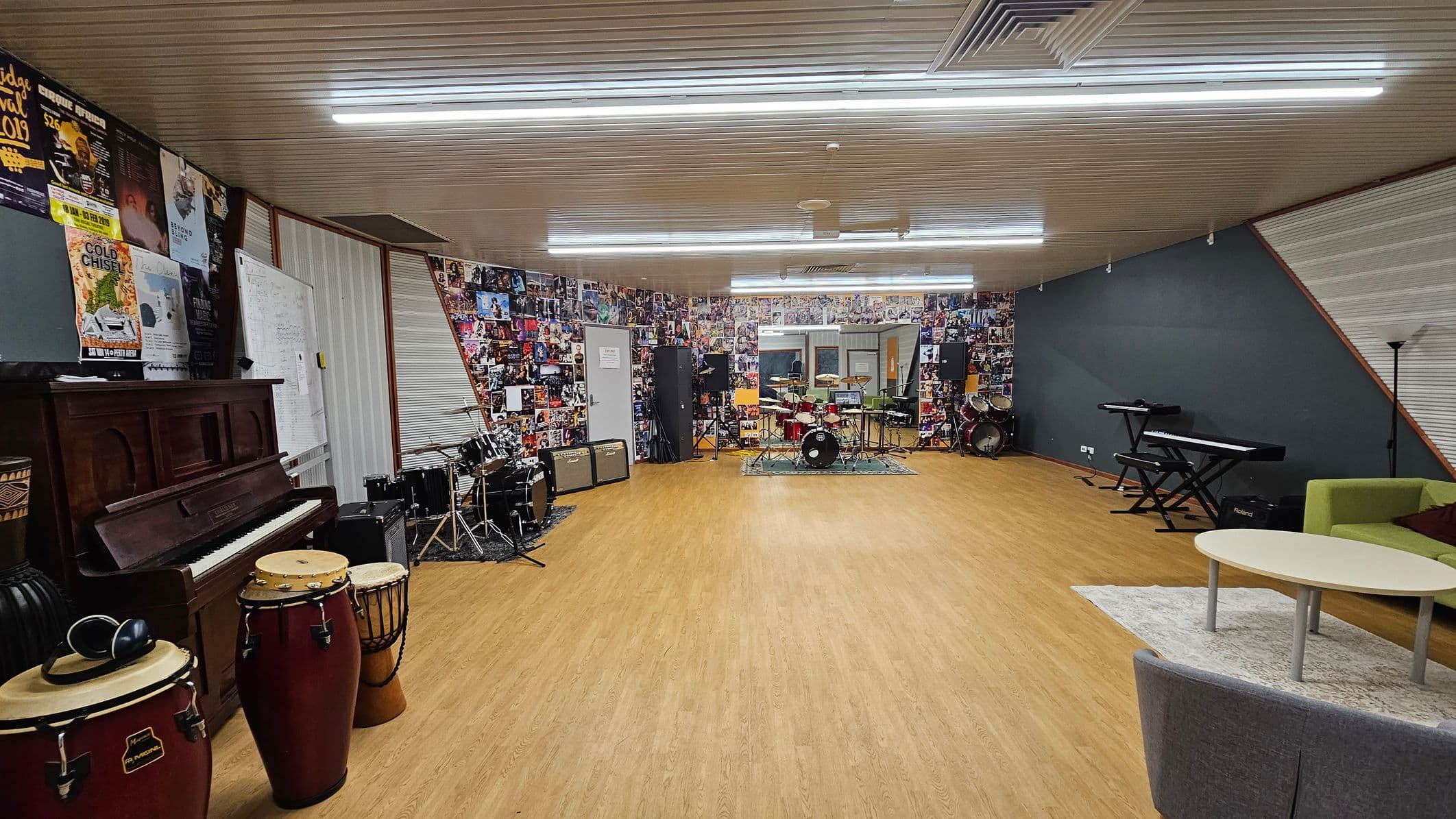 A spacious music room with a wooden floor and a ceiling with fluorescent lights. On the left, there is a piano and a set of conga drums. The back wall is covered with posters and features a drum kit and amplifiers. On the right, there is a keyboard on a stand, a green sofa, and a round table on a rug. The walls are decorated with numerous music-related images and posters.