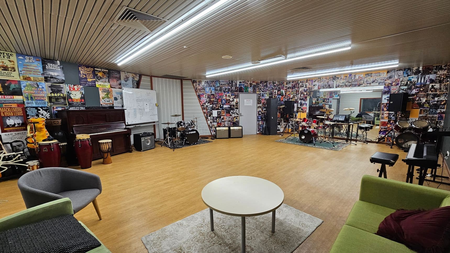 A spacious music room with wooden flooring and a ceiling with fluorescent lights. The left side features a wall covered in posters, a piano, and various percussion instruments. In the centre, there's a round table on a rug, surrounded by chairs and a green sofa. The right side has a wall adorned with photos, multiple drum kits, amplifiers, and a keyboard. A whiteboard is also visible on the left wall.