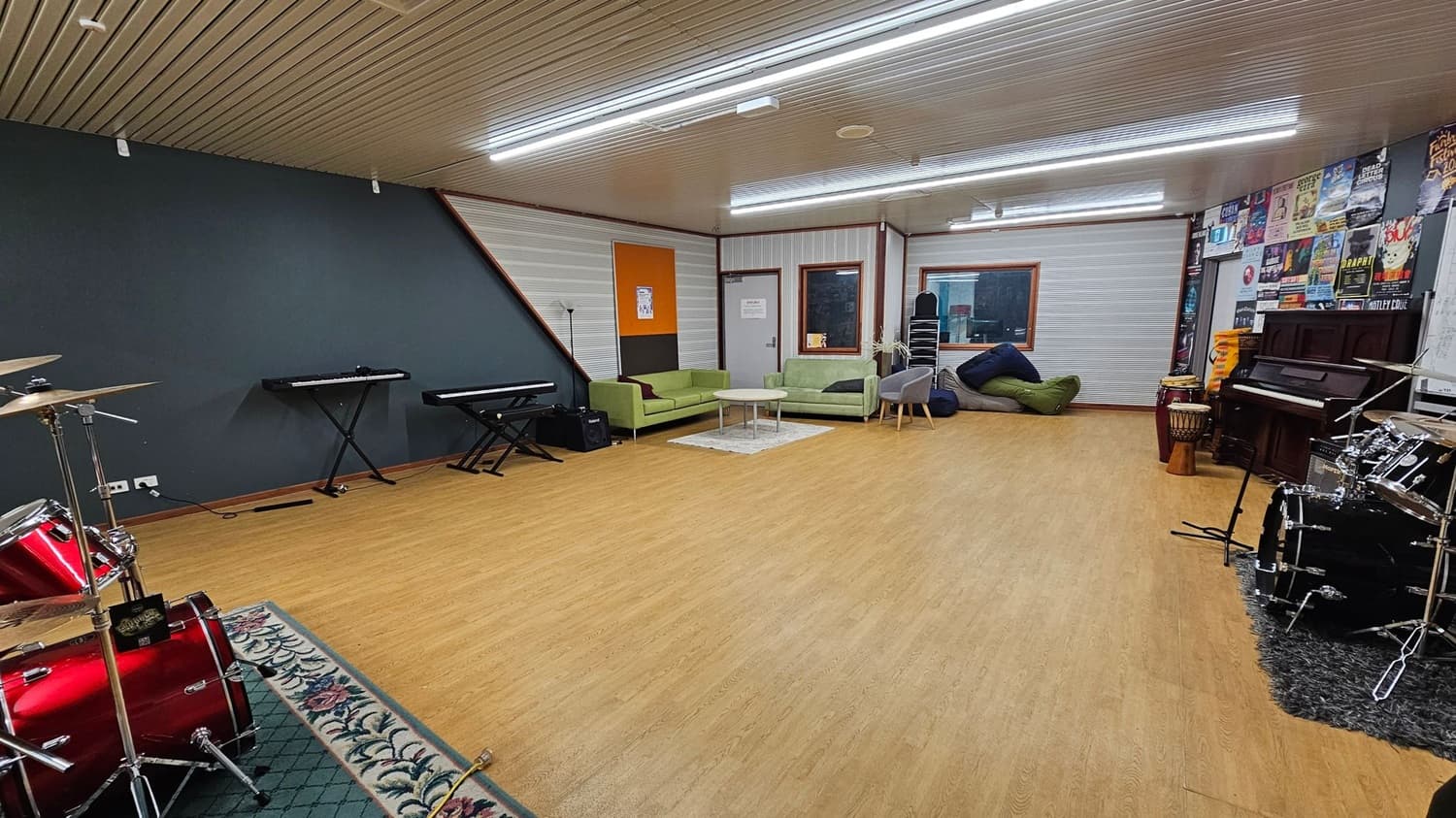 A spacious music room with a wooden floor and a ceiling with fluorescent lights. On the left, there are two electronic keyboards against a dark wall, and a red drum set on a patterned rug. In the centre, green sofas and a small table are placed on a light rug. The right side features a wall with posters, a piano, and additional drums. A window and a door are visible in the background, along with stacked chairs and bean bags.