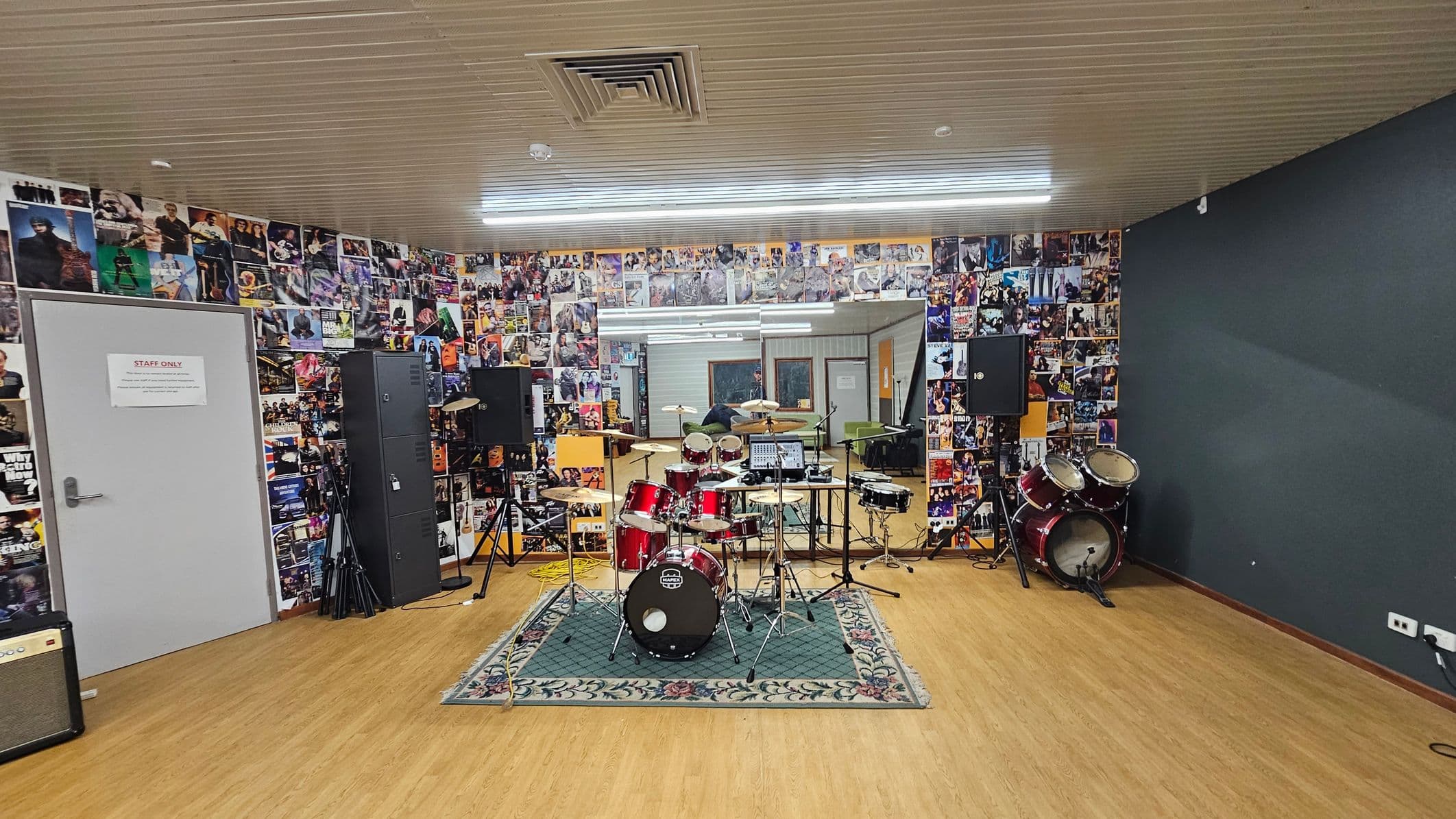 A music rehearsal room with a wooden floor and a ceiling featuring a vent. In the centre, a red drum kit is set on a patterned rug. The back wall is covered with a collage of music posters and a large mirror. To the left, a grey door marked "Staff Only" is visible, alongside speakers and equipment. On the right, additional drums and speakers are present. The room is well-lit with fluorescent lights.