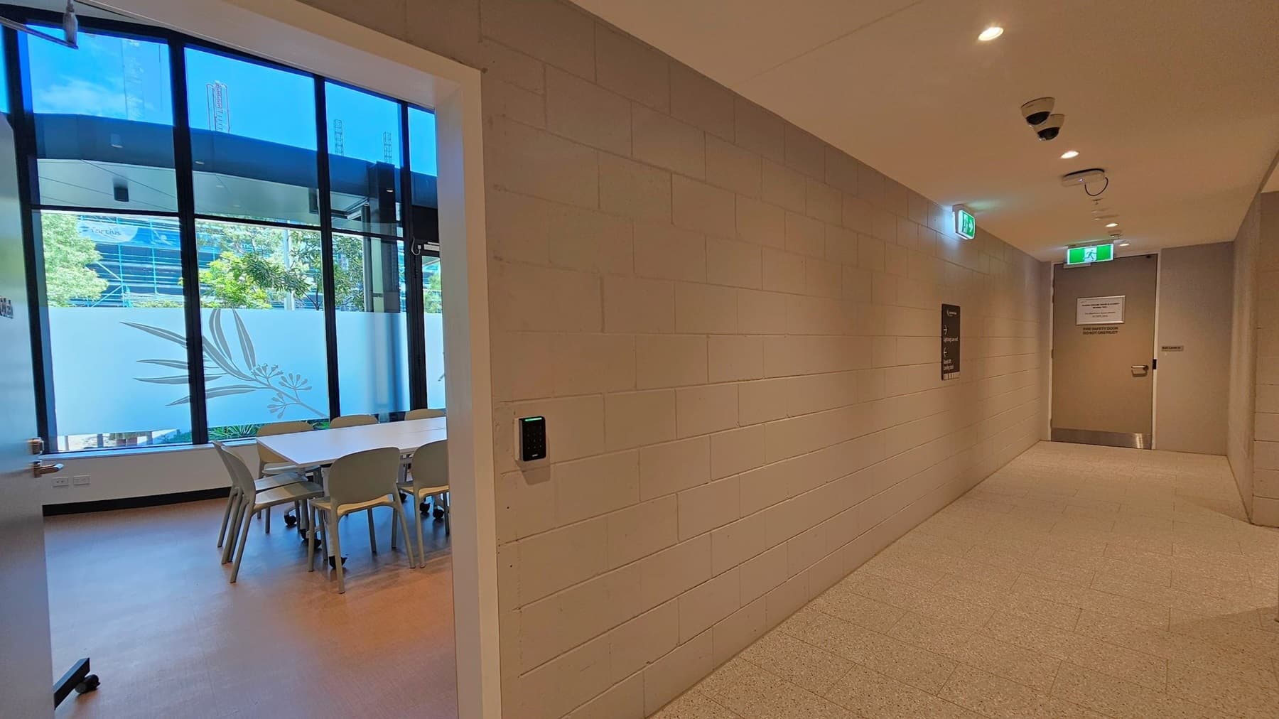 A hallway with a beige tiled floor and a ceiling with recessed lighting. On the right, a brick wall features a security panel and a sign. Two doors are visible, one at the end of the hallway with a sign and an exit light above. On the left, an open door leads to a room with a large window, showing a view of trees and a building. Inside the room, there is a table surrounded by chairs on a wooden floor.