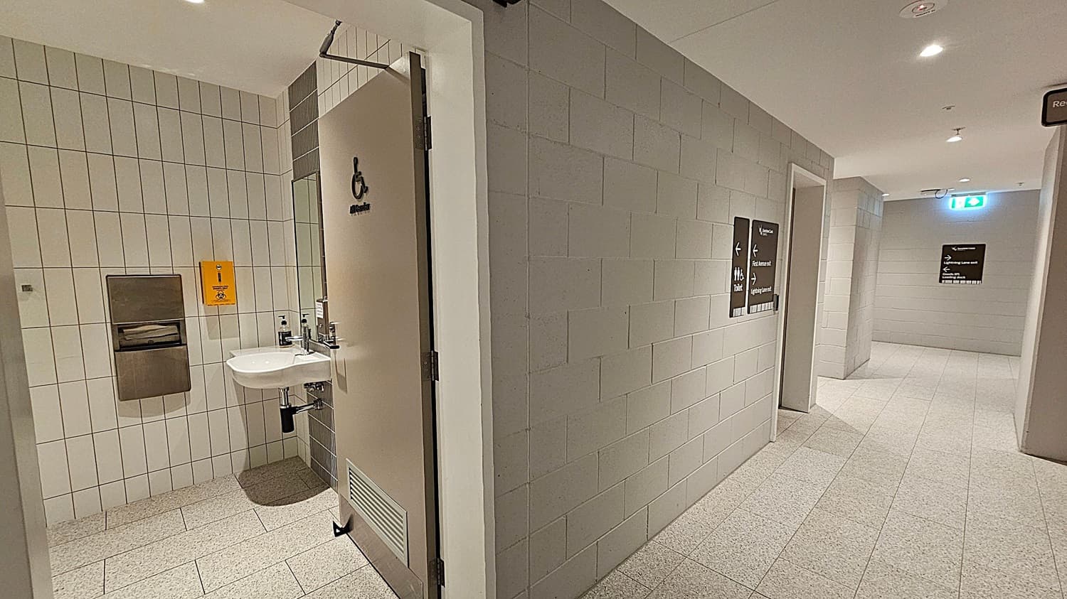 A corridor with light-coloured tiled walls and floor. On the left, an open door leads to an accessible toilet with a sink, soap dispenser, and paper towel holder. A yellow bin is mounted on the wall. On the right, signs indicate directions to toilets and other facilities. The ceiling is white with recessed lighting. An emergency exit sign is visible in the background.