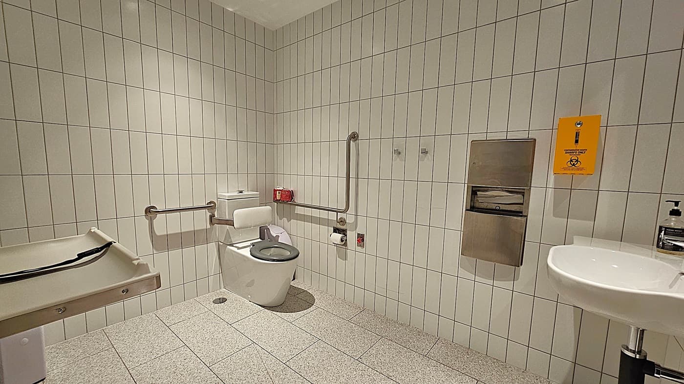 A public accessible toilet with white tiled walls and a speckled grey floor. On the left, there's a baby changing station. In the centre, a toilet with support rails and a green seat cover. Above the toilet, a red box and toilet paper holder are mounted on the wall. On the right, a sink with a soap dispenser is next to a metal paper towel dispenser. A yellow sharps disposal sign is visible above the dispenser. The ceiling is plain white.