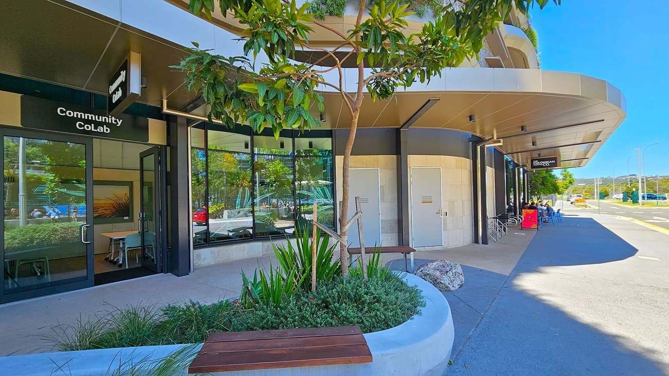 Entrance to a modern building with large glass windows and a curved design. On the left, there's a sculpture of two stylised fish with geometric patterns. The floor is paved with light-coloured tiles. On the right, a wooden bench and a sign for "Altitude Nine" are visible. A potted plant and a small garden area add greenery. The ceiling has a wooden finish with recessed lighting.