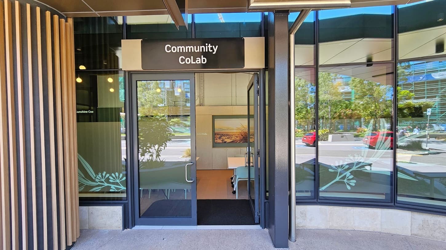 Entrance to a building with a sign reading "Community CoLab" above the door. The door is glass with metal handles, and the interior shows a table and chairs. The left side has vertical wooden slats, while the right side features large glass windows reflecting trees and parked cars outside. Inside, a framed picture is visible on the wall. The floor is concrete, and the ceiling has a dark finish with visible beams.