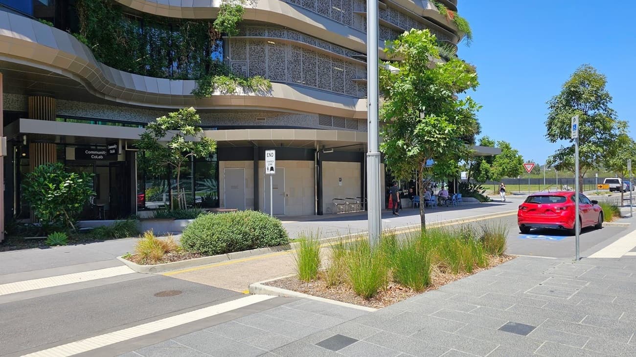 On the left, a modern building with curved architecture and greenery on the facade. In front, a sign reads "Community CoLab". The middle features a pathway with bushes and tall grass. On the right, a red car is parked in a designated space, with a parking sign nearby. A few people are seated at outdoor tables under the building's overhang. Trees line the street, and a "Give Way" sign is visible in the background. The pavement is grey with a textured pattern.