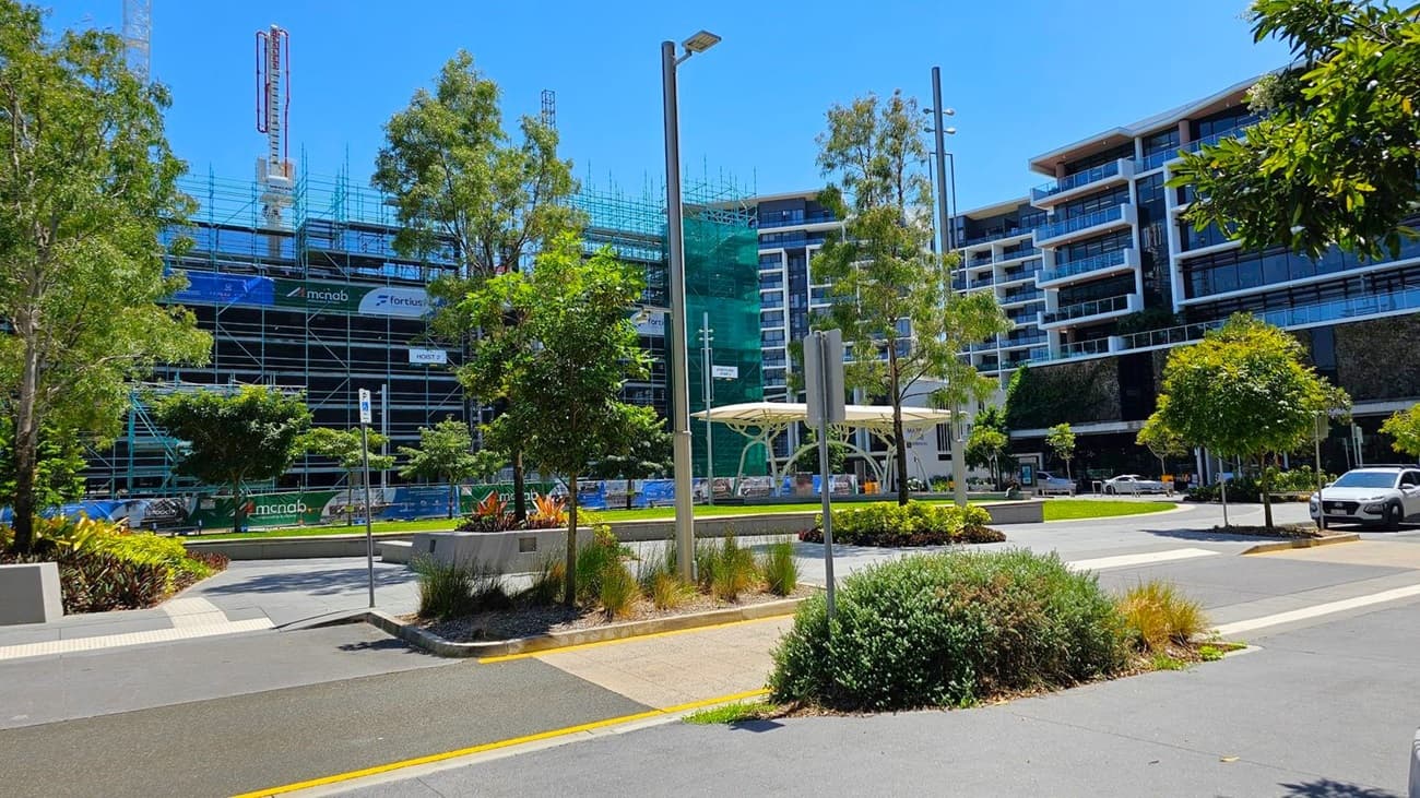 A construction site on the left with scaffolding and cranes, surrounded by trees. In the centre, a small park area with a modern white canopy structure and landscaped greenery. On the right, a multi-storey building with large windows and balconies. A car is parked on the street, and there are street lamps and signs. The ground is paved with a mix of concrete and asphalt.