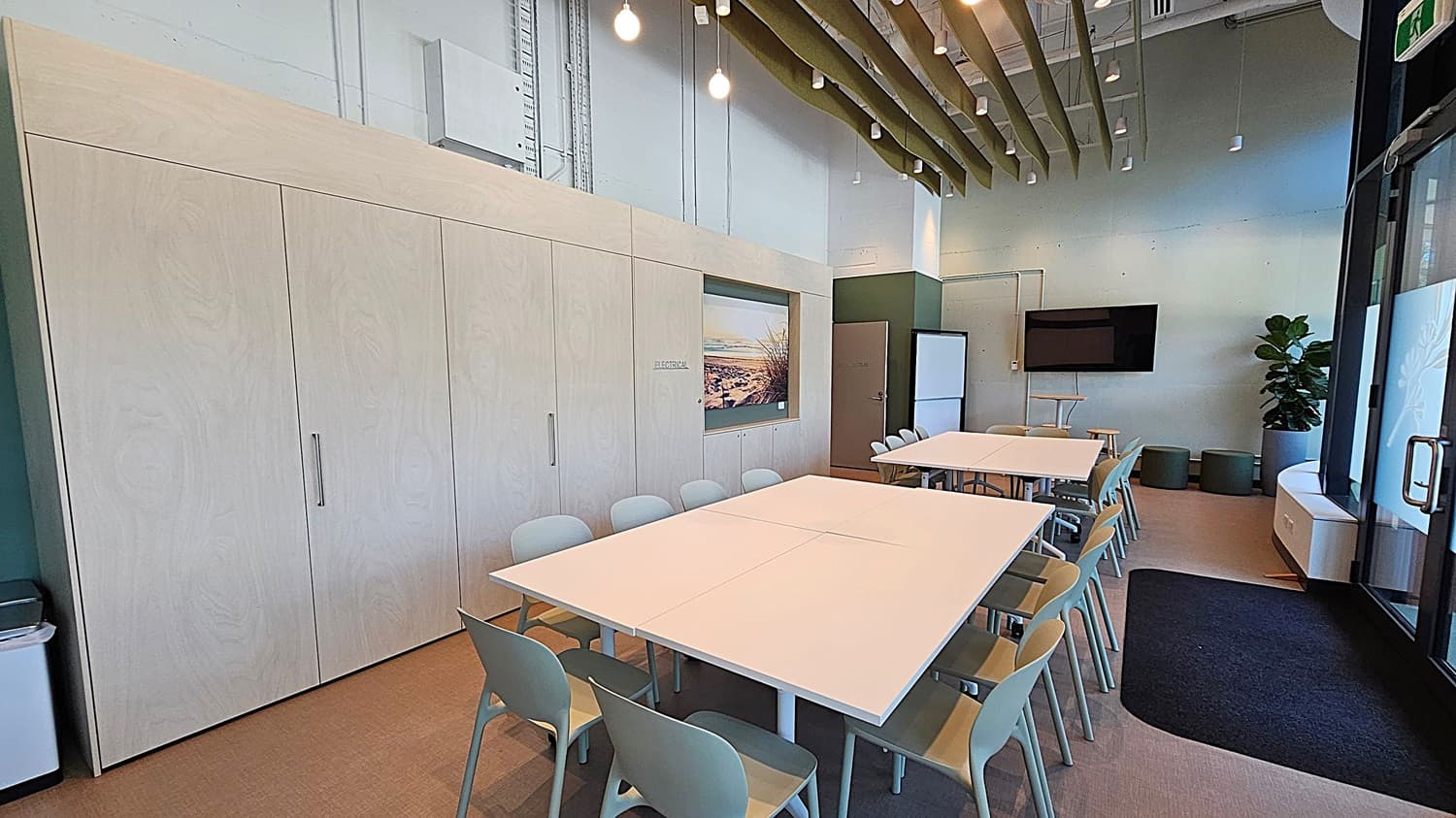 A modern office space with a kitchenette on the left, featuring white cabinets, a sink, and built-in appliances. In the centre, there's a large framed photograph of a beach scene. To the right, an open door leads to another room. In the foreground, there are white tables with chairs on wheels. The walls are a mix of white and green, and the ceiling is high with visible pipes and lighting fixtures.