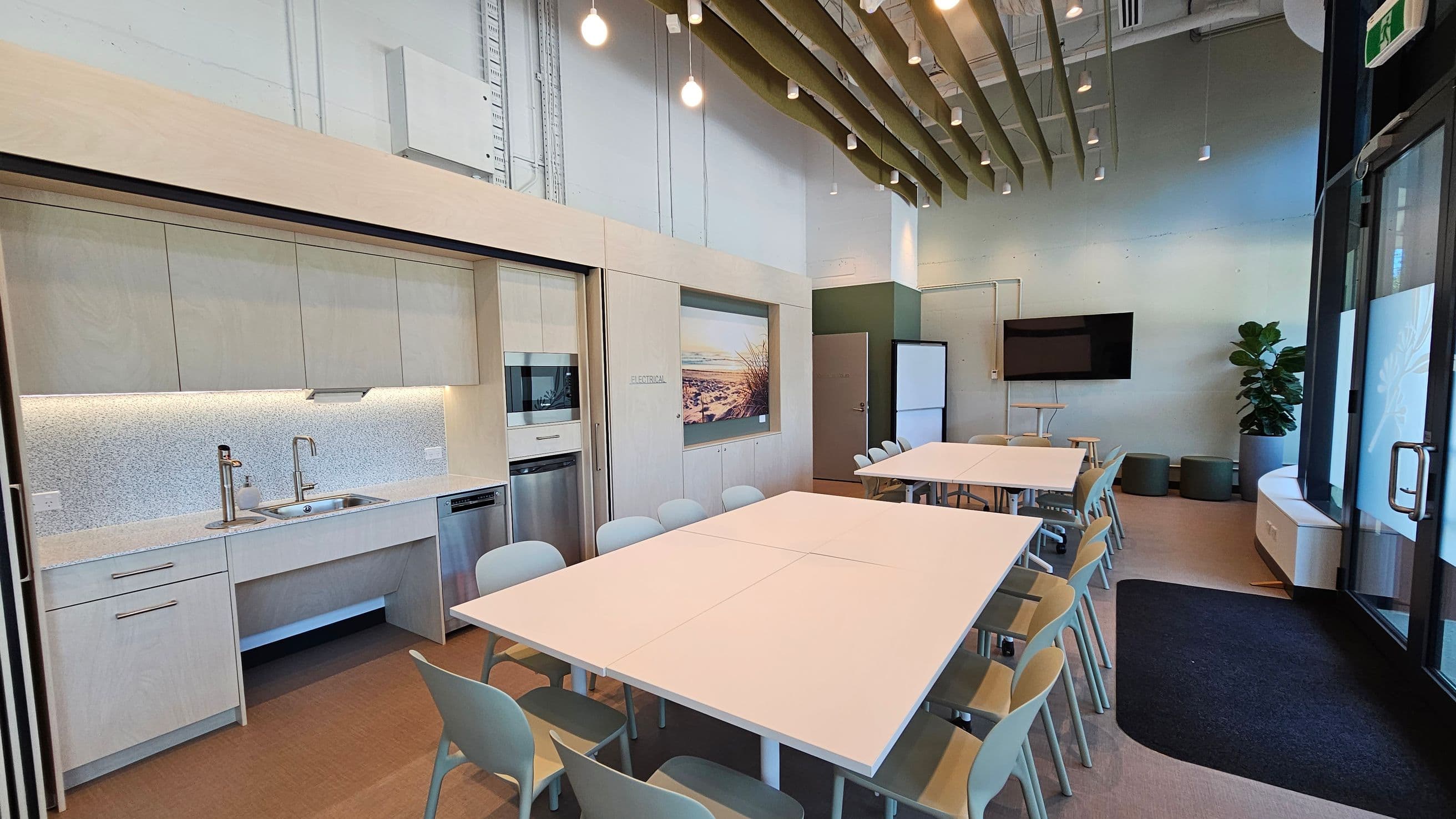 A modern kitchen and meeting area with light wood cabinetry on the left, featuring a sink, microwave, and dishwasher. In the centre, there are two large white tables surrounded by light grey chairs. The right side has a wall-mounted TV, a plant, and a glass door. The ceiling has exposed beams and hanging lights, and the floor is a mix of wood and carpet. A large photograph is displayed on the wall above the cabinets.
