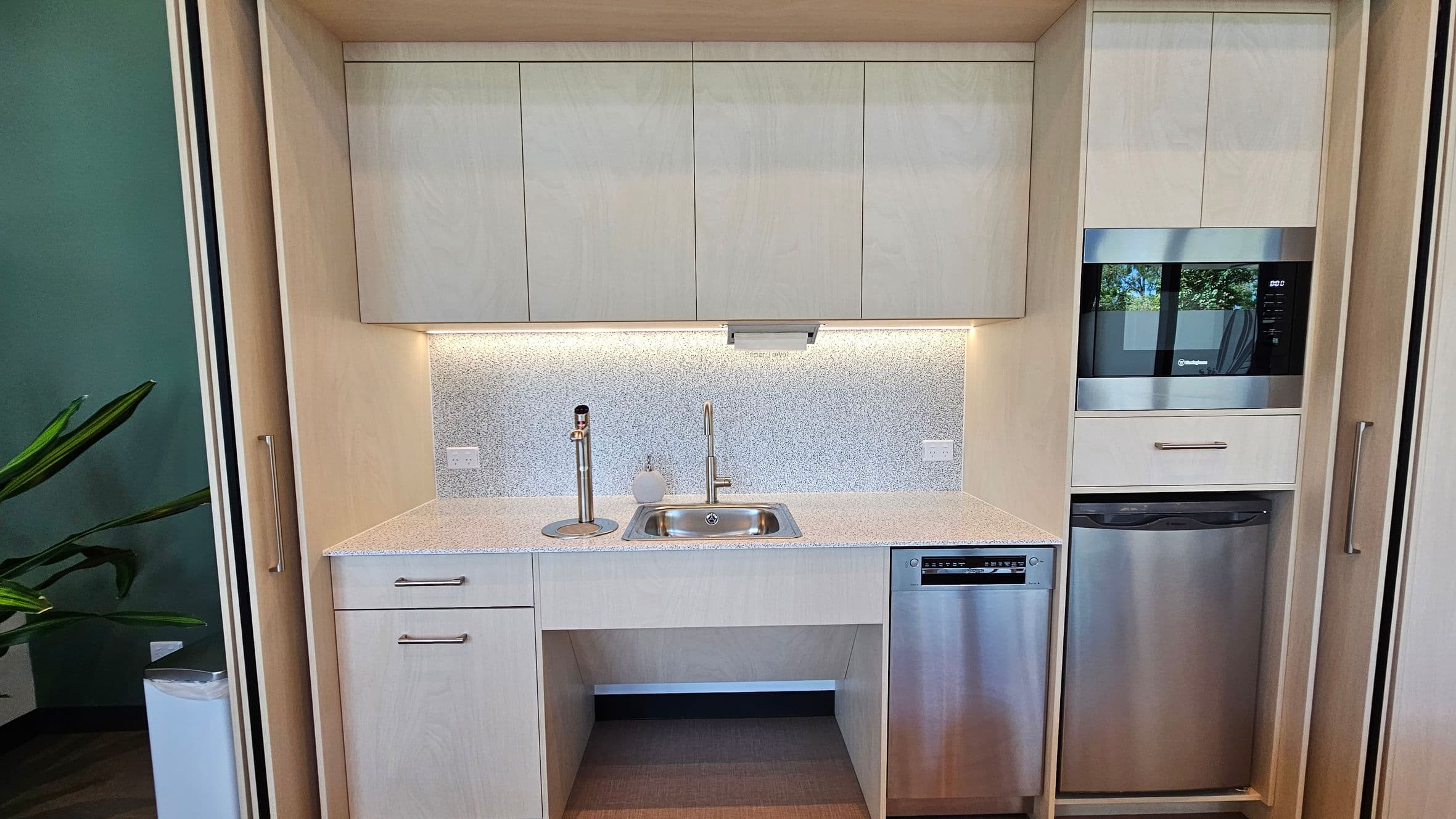 A compact kitchen area with light-coloured cabinetry. In the centre, there's a small sink with a modern tap, and a soap dispenser on the left. Above the sink are wall-mounted cabinets. To the right, a built-in microwave sits above a stainless steel dishwasher. The backsplash is speckled, and there's under-cabinet lighting. On the left, a glimpse of a green wall and a plant is visible. The floor is a light wood tone.