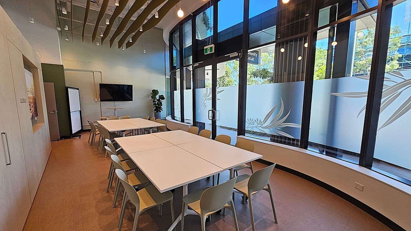 A modern meeting room with a large rectangular table in the centre surrounded by beige chairs. On the left, there is a wall with a door and a mounted flat-screen TV. The right side features large windows with frosted glass designs, allowing natural light to enter. A potted plant is near the corner. The ceiling has hanging lights and decorative wooden slats. The floor is carpeted in a light brown tone.