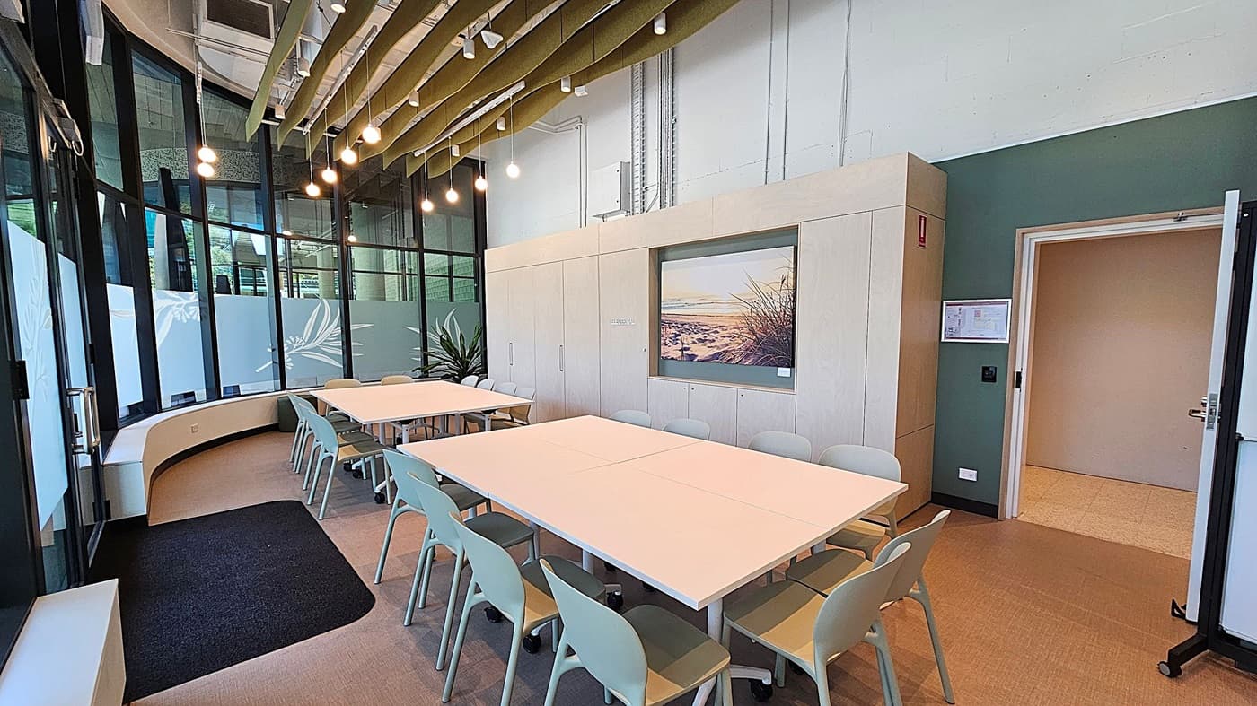 A modern meeting room with a large rectangular table surrounded by light grey chairs. On the left, there are tall windows with frosted glass panels and a curved bench. The ceiling features exposed beams and hanging lights. In the centre, a wall with a landscape picture and storage cabinets. On the right, an open door leads to another room, and a green wall with a mounted display. The floor is carpeted in a neutral tone.