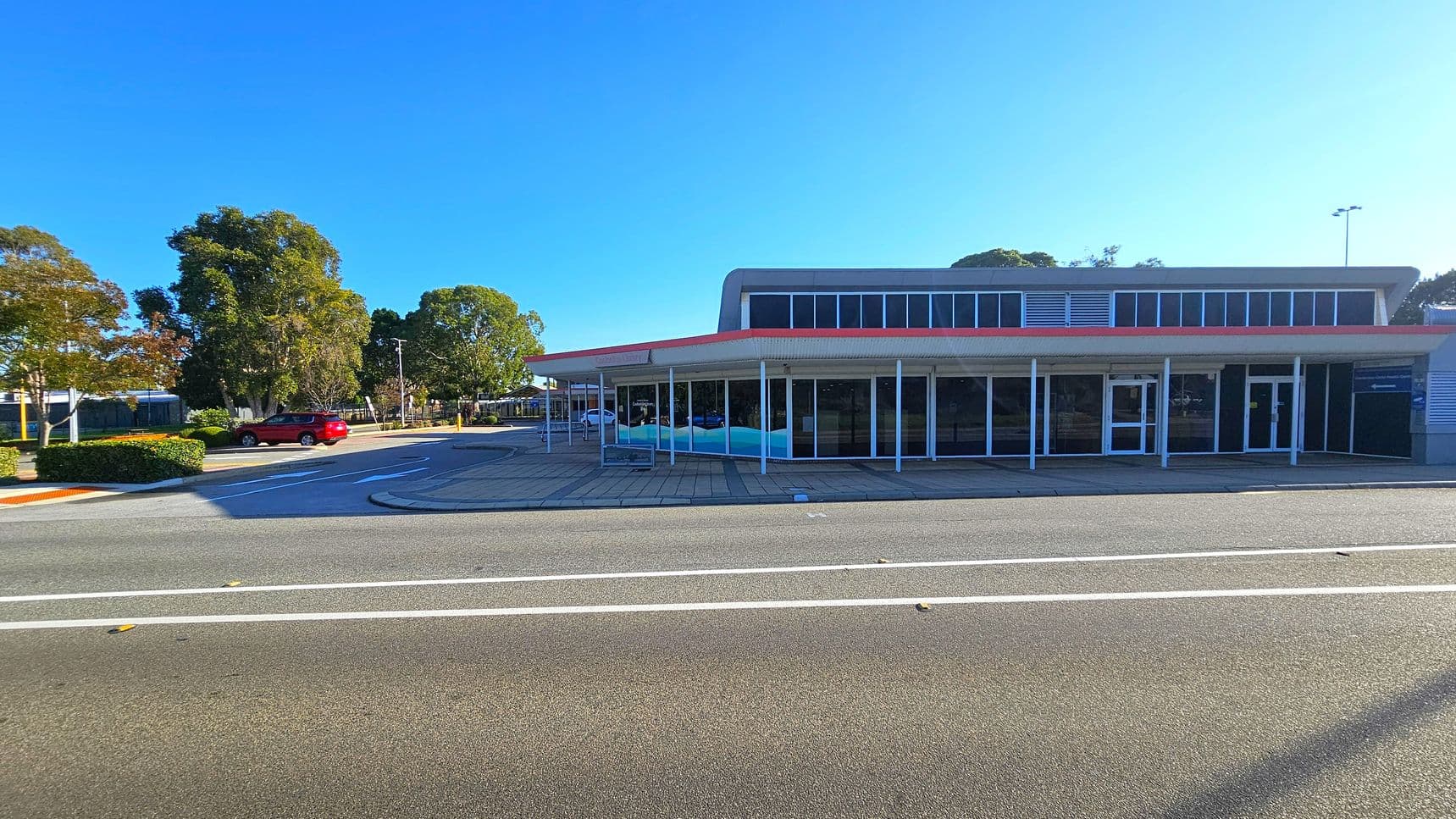 A single-storey building with large glass windows and a red trim roof is on the right. The pavement in front is tiled. On the left, a red car is parked near some trees and bushes. The road in the foreground has white lane markings. The background features more trees and a clear blue sky.