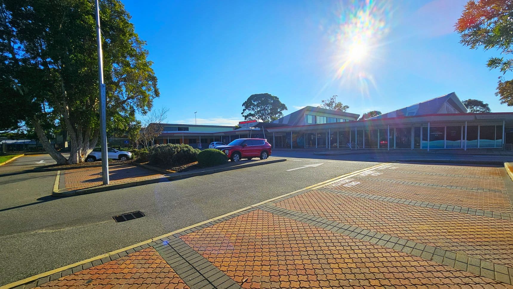 A paved area with interlocking bricks in the foreground. To the left, a large tree stands next to a metal pole, with a white car parked nearby. In the centre, a red SUV is parked near a low bush. The background features a single-storey building with large windows and a sloped roof. The sun is shining brightly in the sky, casting light across the scene.