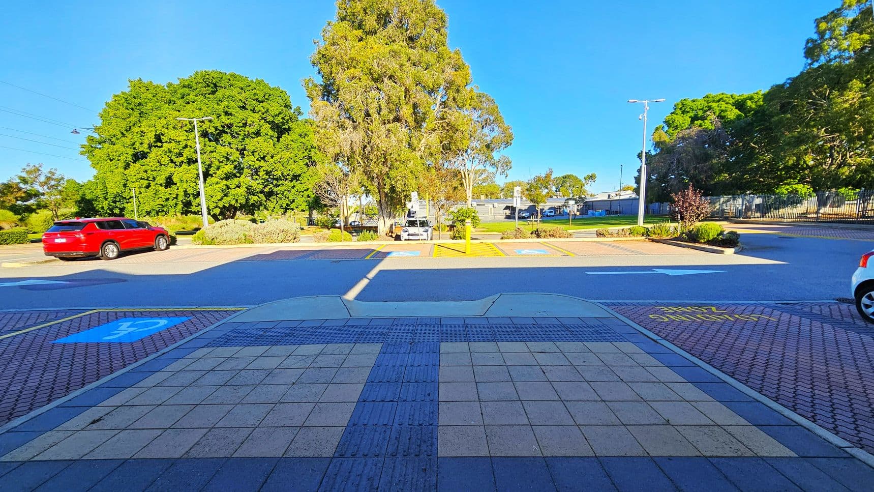 A parking area with a red car on the left and a white car partially visible on the right. In the centre, there are parking spaces marked with blue and yellow lines, indicating disabled parking. Large green trees are in the background, with a few streetlights and a building partially visible. The foreground has a tiled pavement with a textured strip, and the words "SLOW" painted in yellow on the ground.