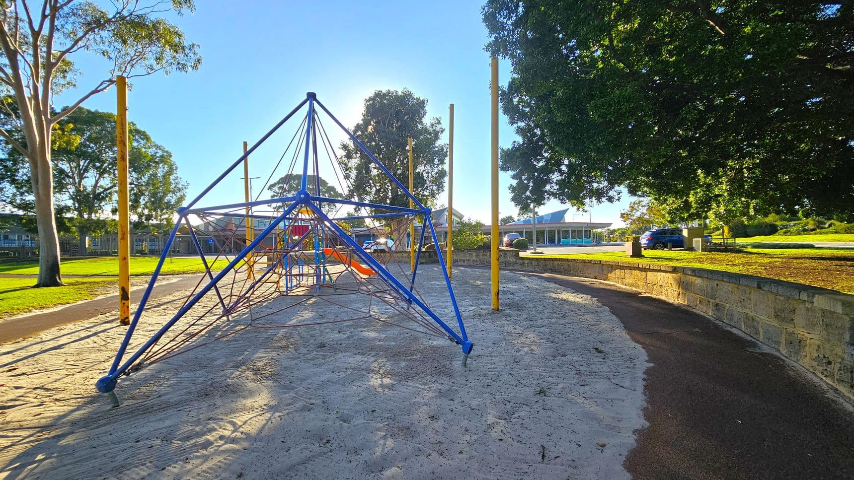A playground with a blue climbing structure made of ropes and poles on a sandy surface. To the left, there are tall trees with green foliage. On the right, a stone wall borders a paved path, with a large tree providing shade. In the background, a building with a blue roof and a parked car are visible. The area is well-lit, suggesting a sunny day.