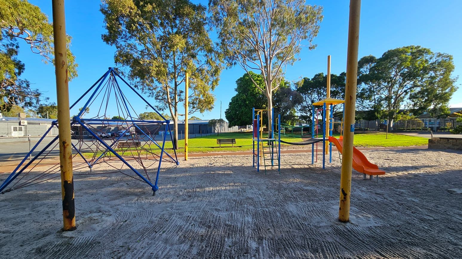 A playground with a sandy surface. On the left, there's a blue climbing structure made of ropes and poles. On the right, a playset with a slide and climbing bars, featuring orange and yellow elements. In the background, there are several large trees and a grassy area with a bench. A building with a grey exterior is visible in the distance.