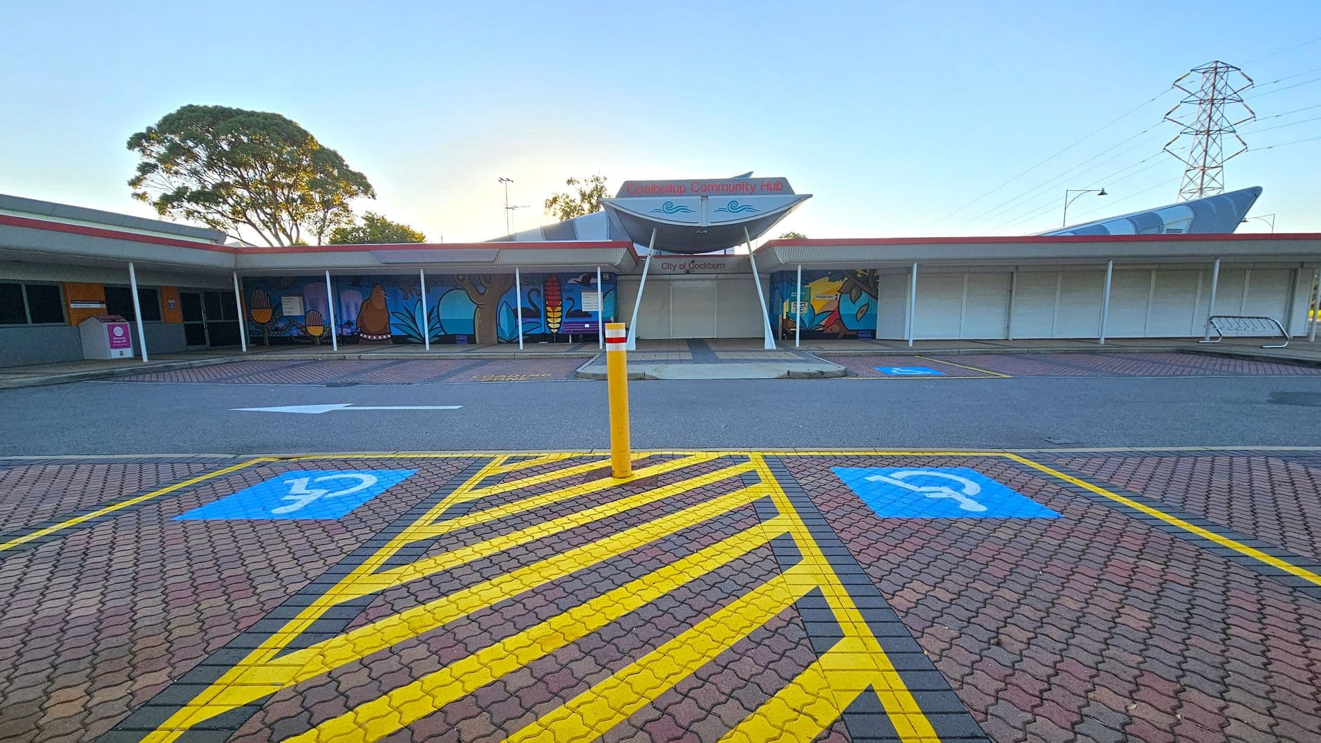 A community hub with a wide entrance and a canopy displaying the name "City of Cockburn." The building features colourful murals on the walls. In the foreground, there are marked parking spaces with blue disability symbols and yellow striped lines. A yellow bollard stands in the centre. The ground is paved with interlocking bricks. To the left, a large tree is visible, and to the right, there is a tall electricity pylon.