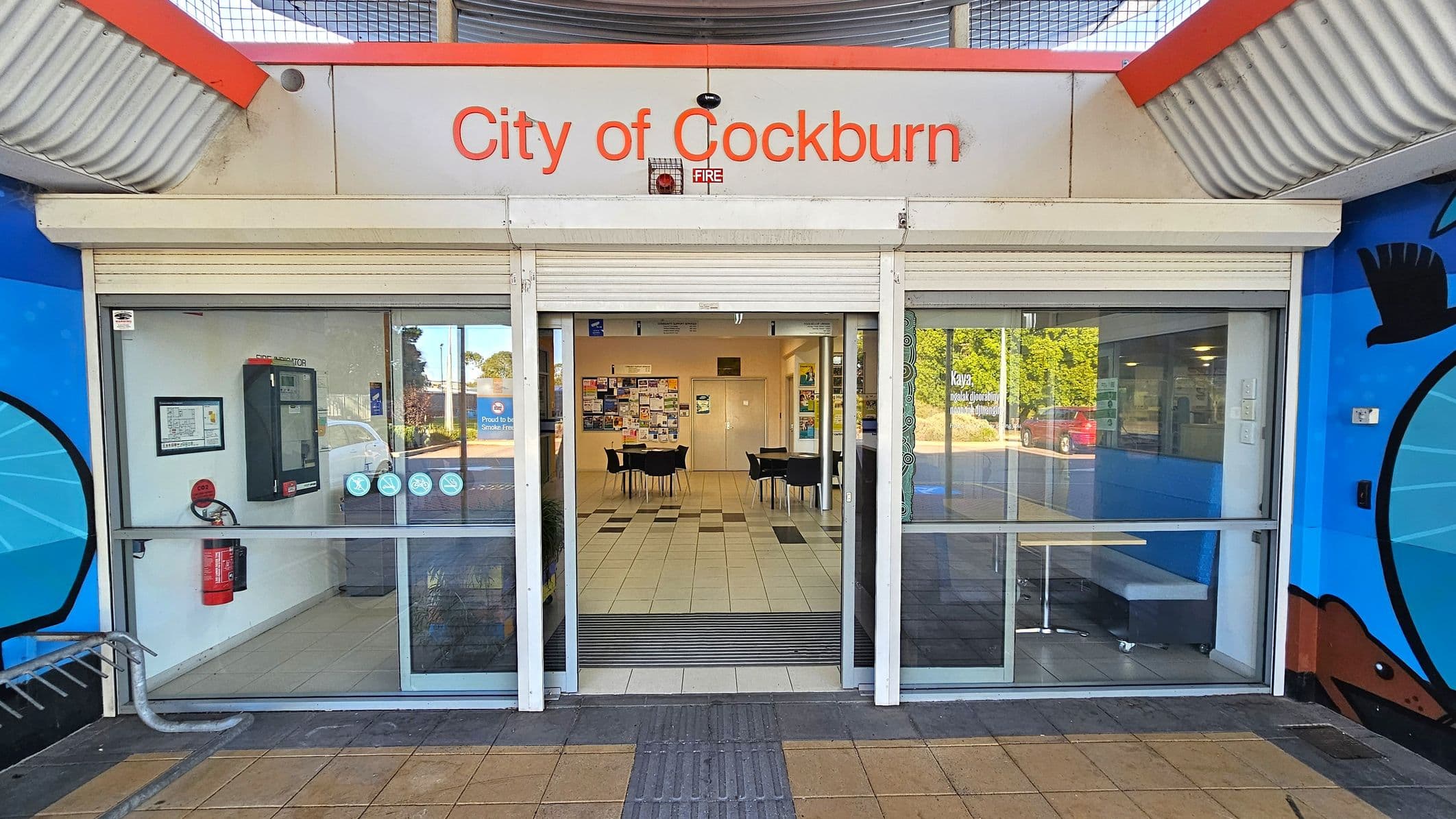 Entrance to a building with a sign reading "City of Cockburn" above glass sliding doors. The left side features a fire extinguisher and a wall-mounted device. Inside, there are tables and chairs on a tiled floor, with notice boards on the far wall. The right side shows a window with a view of a red car and greenery outside. The exterior walls are painted with blue artwork, and the ceiling has a corrugated metal design.