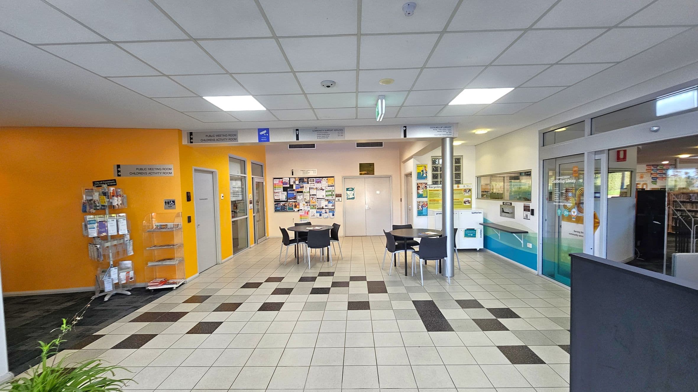 A library entrance area with a tiled floor featuring a pattern of light and dark squares. On the left, there's a bright orange wall with a brochure stand. In the centre, several round tables with black chairs are arranged. The back wall has a bulletin board filled with notices. On the right, glass doors lead into the library, with a sign above them. The ceiling is white with recessed lighting.
