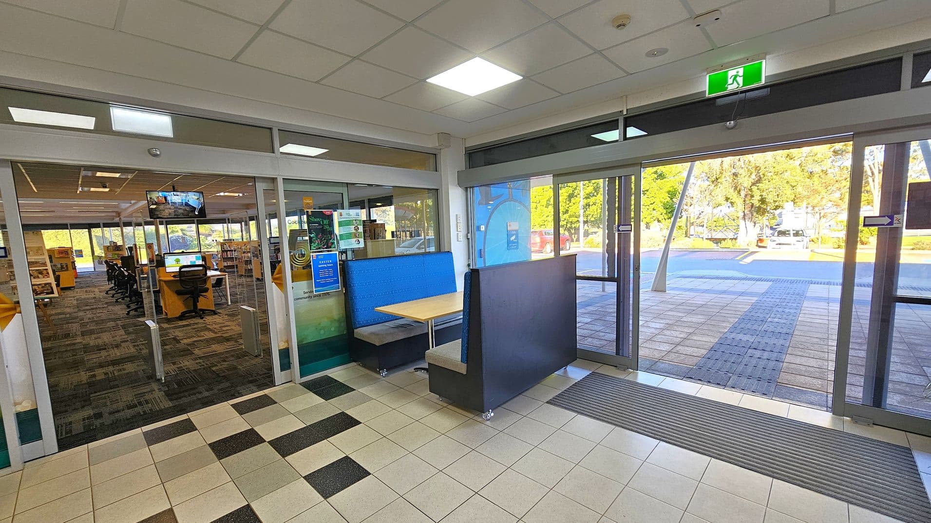 A library entrance with glass doors on the left leading to an interior with bookshelves and computer stations. A blue booth with a wooden table is positioned near the entrance. On the right, large glass doors open to an outdoor area with trees and a parked car visible. The floor is tiled, and the ceiling has recessed lighting and an illuminated exit sign.