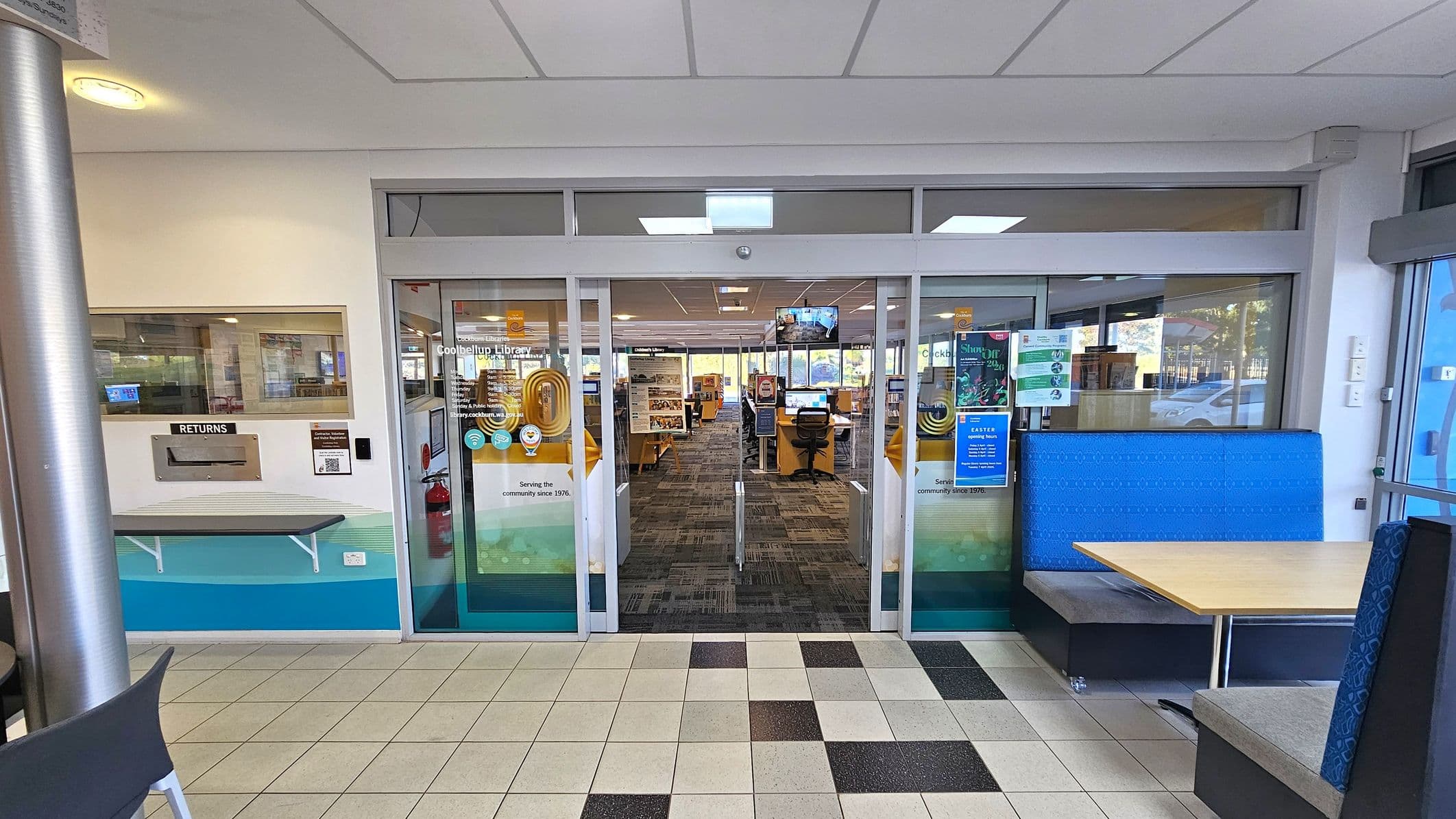 A library entrance with glass sliding doors in the centre, displaying various notices and signs. To the left, there's a book return slot with a small shelf beneath it. On the right, a seating area with a blue upholstered booth and a wooden table. The floor is tiled with a mix of light and dark squares, and the ceiling has recessed lighting. Inside the library, bookshelves and computer stations are visible.