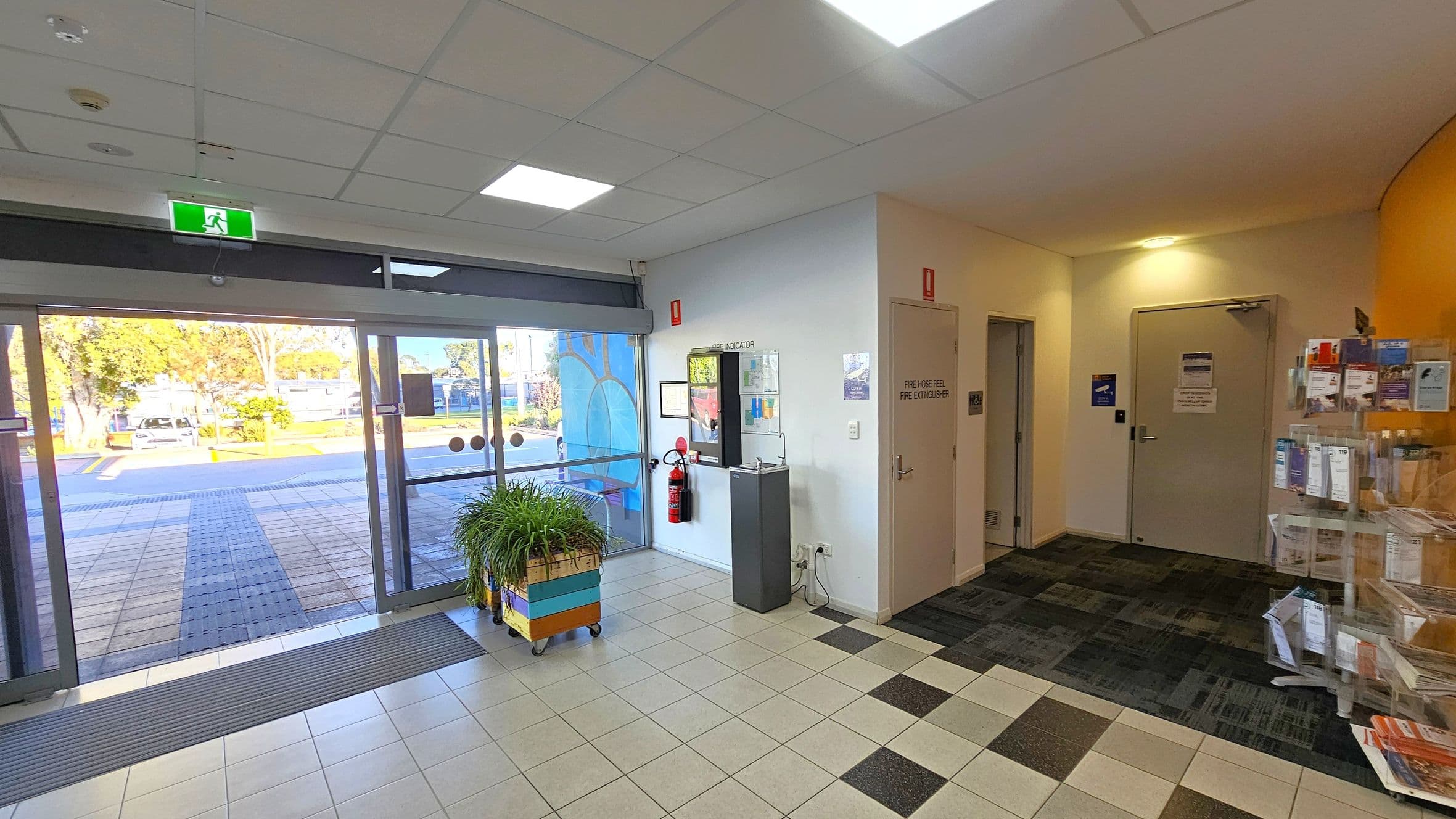 A spacious indoor area with a tiled floor. On the left, there are large glass doors leading outside, with a green exit sign above. A planter with green plants is near the entrance. On the right, there are two doors, one marked for fire equipment, and a wall-mounted brochure rack filled with pamphlets. The ceiling is fitted with square lights, and the walls are white. Outside, trees and a parked car are visible through the glass doors.