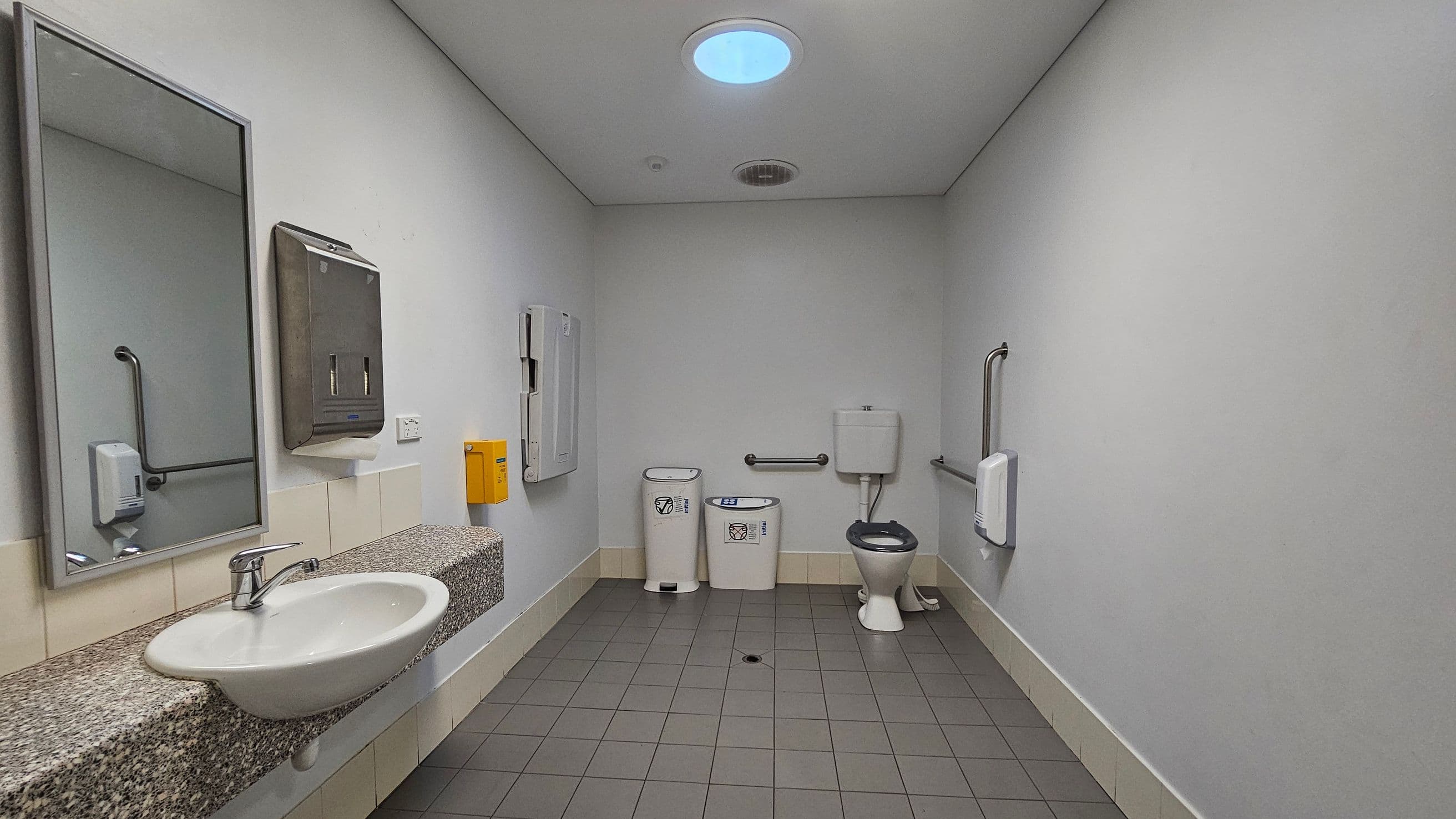 A spacious public restroom with grey tiled flooring and a white ceiling featuring a circular skylight. On the left, a granite countertop with a white sink, a large mirror, and a soap dispenser. A paper towel dispenser and a yellow bin are mounted on the wall. In the centre, two waste bins are placed near a toilet with a support rail. The walls are light grey with a tiled skirting. A baby changing station is mounted on the wall near the toilet.