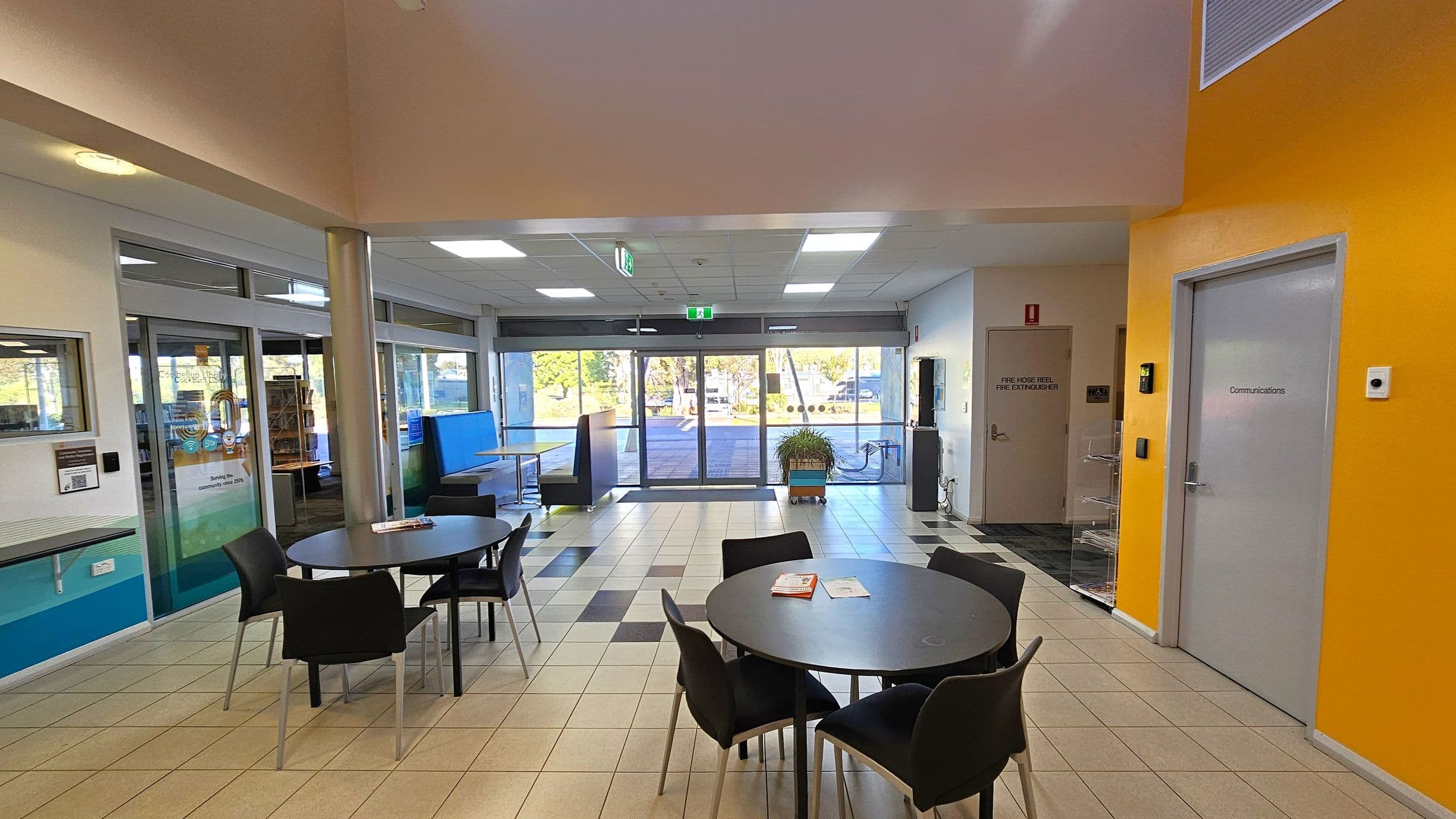 A spacious indoor area with a tiled floor and a ceiling with recessed lighting. In the foreground, there are round tables with black chairs. To the left, glass doors lead to another room with visible bookshelves. The background features large glass doors opening to an outdoor area. On the right, a bright orange wall has a door labelled "Communications" and another door marked for fire safety equipment. A potted plant is near the glass doors.
