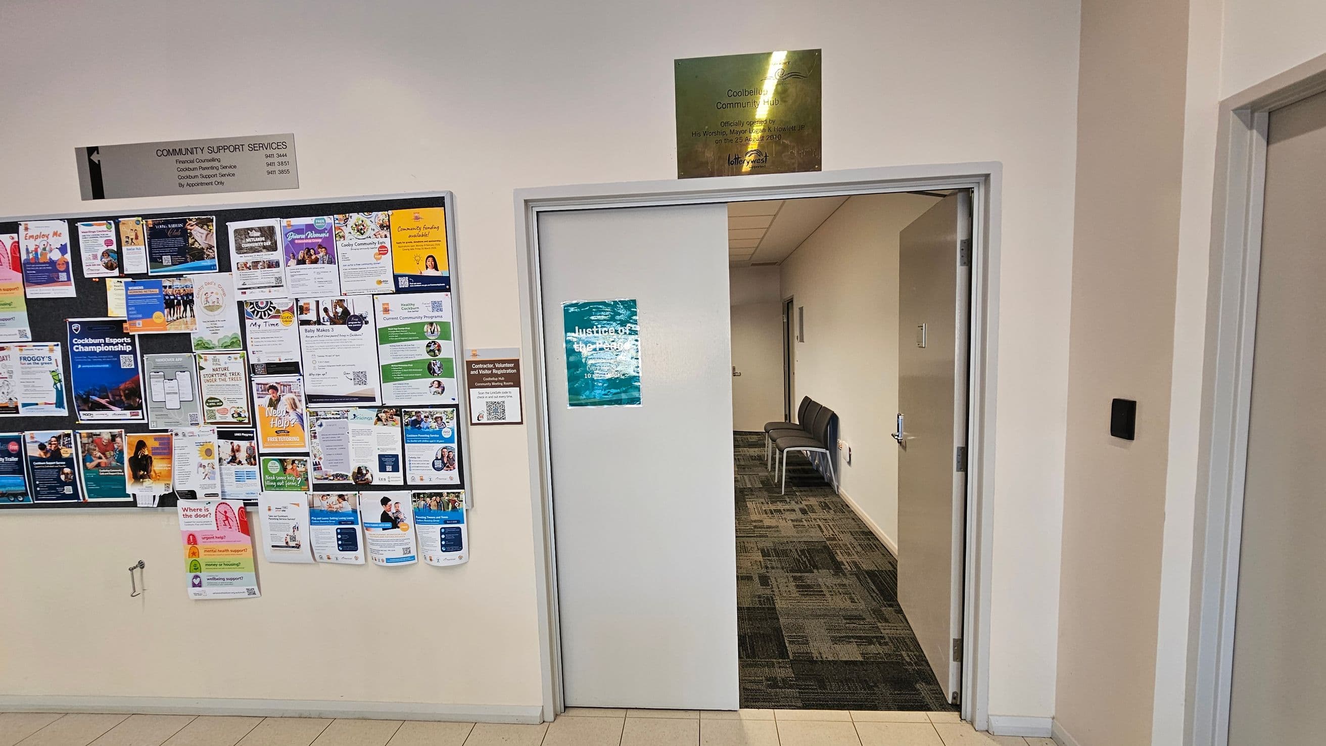 A hallway with a bulletin board on the left displaying numerous colourful flyers and notices. To the right, a partially open door leads to a corridor with a patterned carpet and a couple of chairs against the wall. Above the door, a plaque is mounted on the wall. The walls are light-coloured, and the floor is tiled.
