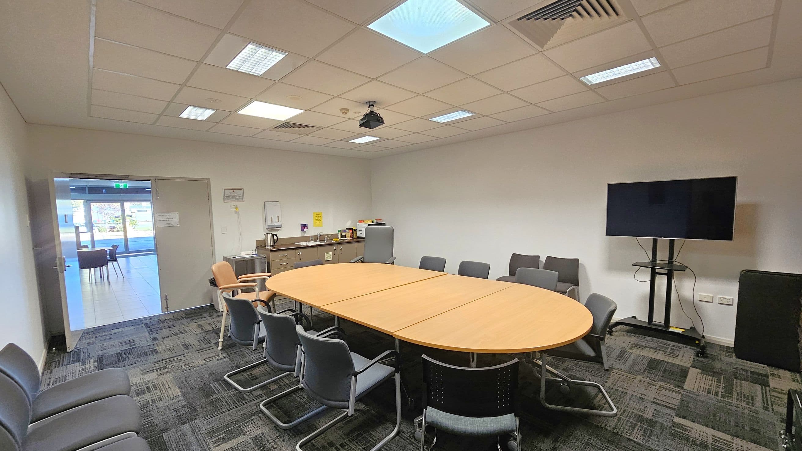 A conference room with a large oval wooden table in the centre surrounded by grey chairs. On the right, a large flat-screen TV is mounted on a stand. The left side has a door leading to a hallway with visible chairs and tables. At the back, a cabinet holds a kettle and various items. The ceiling is fitted with fluorescent lights and a projector. The floor is carpeted with a patterned design.