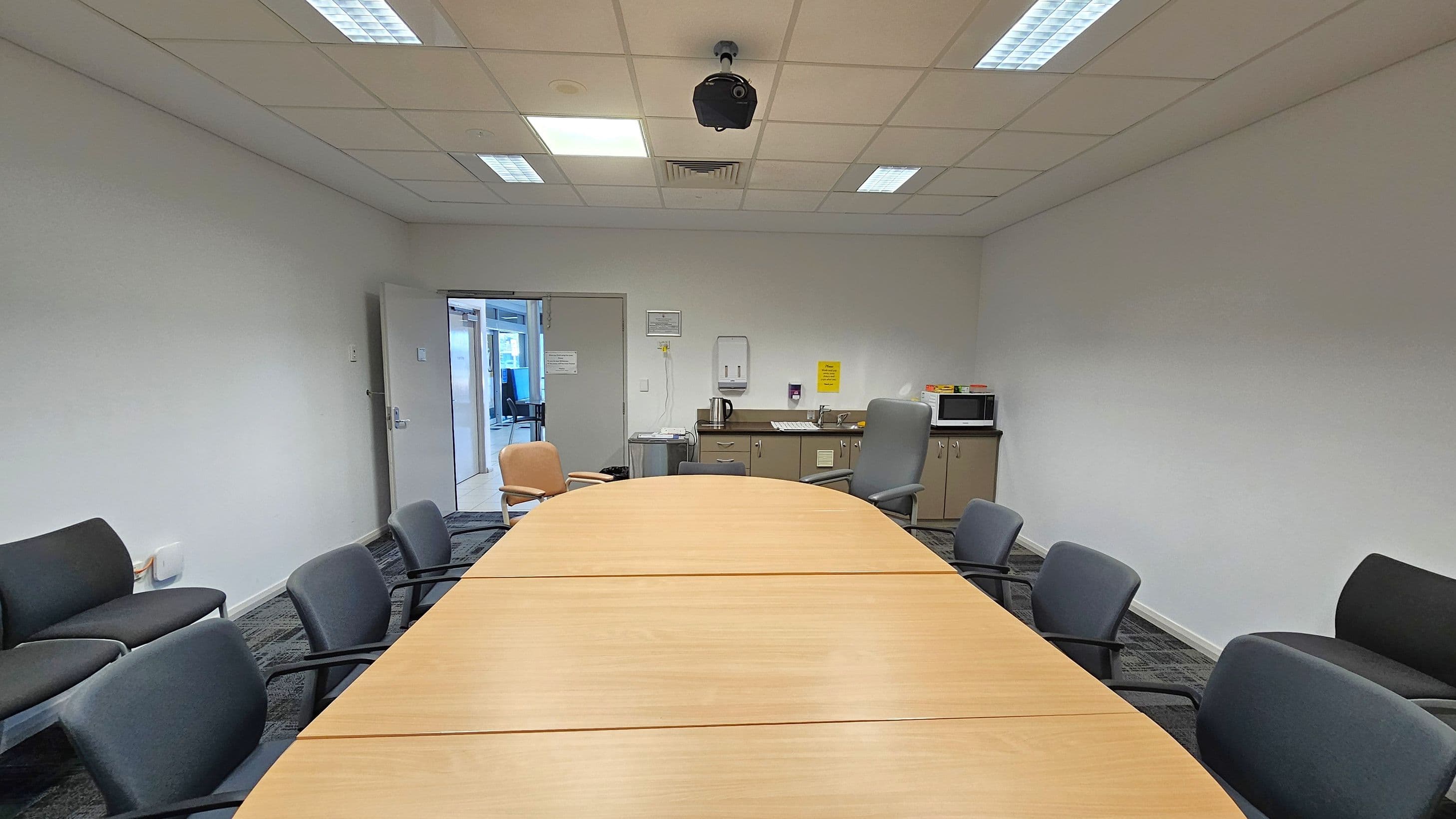A conference room with a long wooden table in the centre surrounded by black chairs. On the left, there are additional chairs against the wall. At the back, a door is open, leading to another room. On the right, a kitchenette area with a microwave, kettle, and sink is visible. The ceiling has fluorescent lights and a mounted camera. The floor is carpeted in a dark pattern.