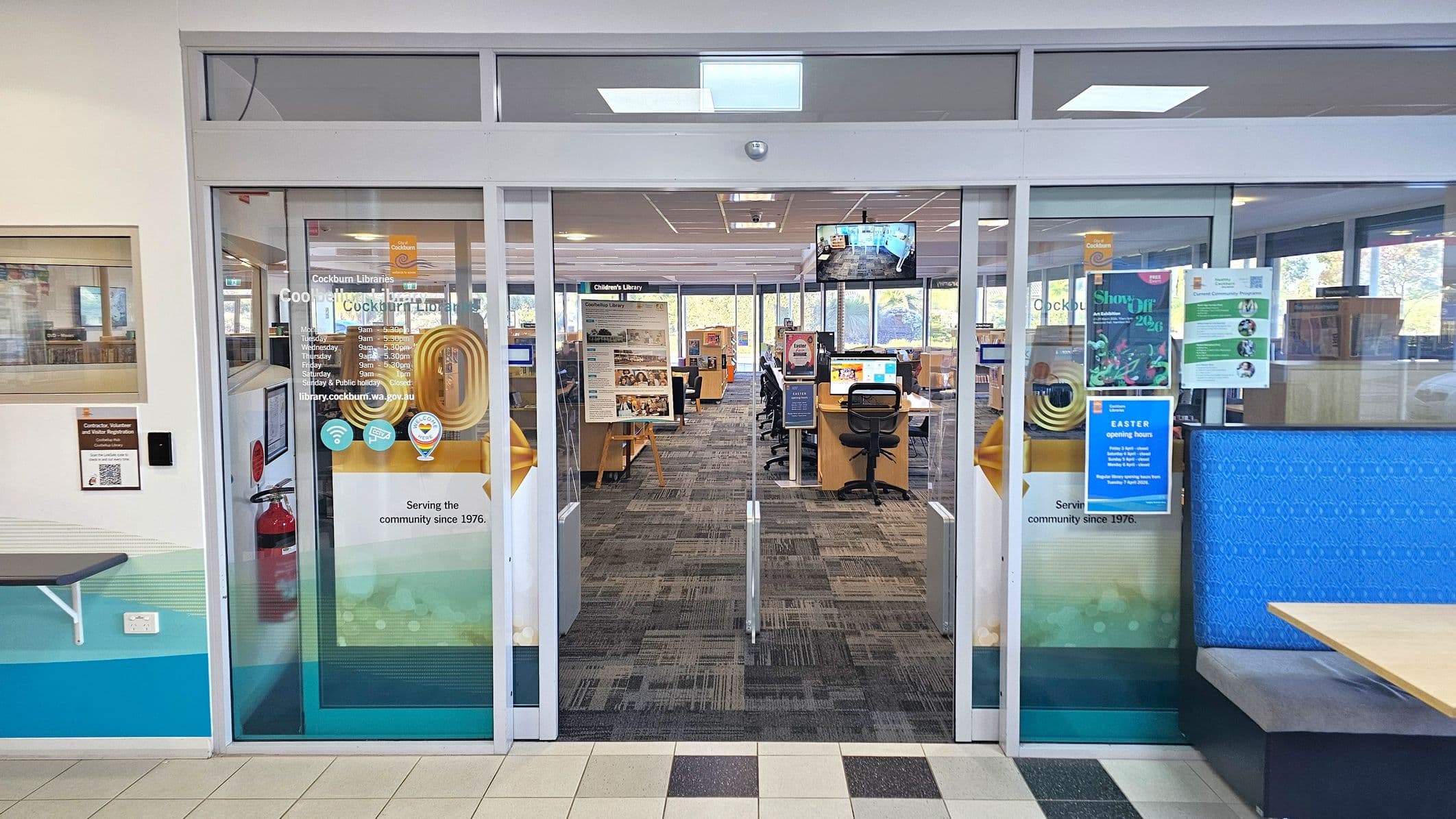 Glass doors lead into a library with a patterned carpet. On the left, a fire extinguisher is mounted on the wall, and a small shelf is visible. The doors display various signs and posters, including opening hours and event information. Inside, there are computer stations with chairs and a visible sign for the Children's Library. The ceiling has recessed lighting, and large windows in the background allow natural light to enter.