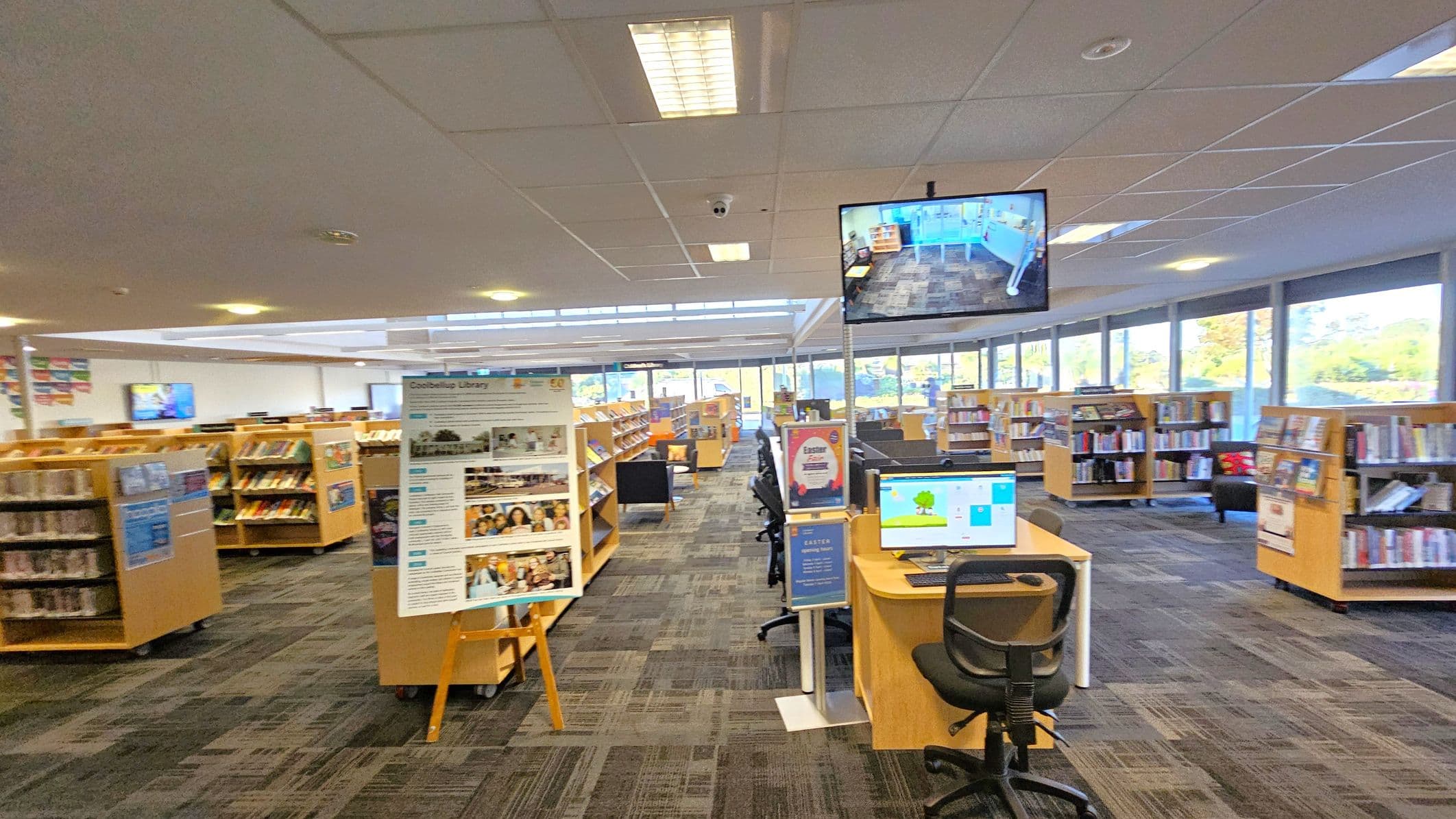 A spacious library interior with rows of wooden bookshelves filled with books. In the foreground, a computer station with a chair and a monitor displaying a colourful screen. A large poster on an easel stands nearby. The ceiling is fitted with fluorescent lights and a security camera. A television screen hangs from the ceiling showing a live feed of the library. Large windows in the background allow natural light to illuminate the space, and the carpet has a patterned design.