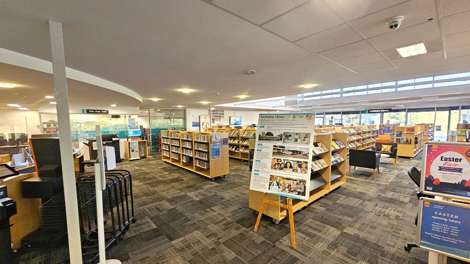 A library interior with a patterned carpet floor and a ceiling with recessed lighting. In the centre, there are wooden bookshelves filled with books. A large informational board on an easel is visible. To the right, a black armchair and a sign about an Easter fair are present. On the left, a print and copy station is visible near a glass-walled area. Large windows in the background allow natural light to enter.