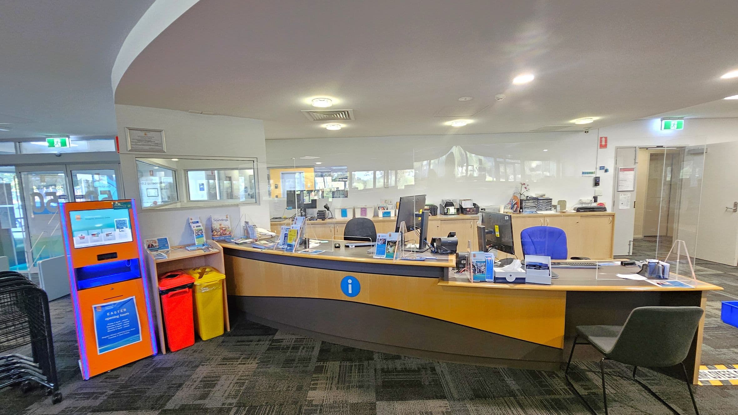 A reception area with a curved wooden desk in the centre, featuring computers, brochures, and a blue chair. To the left, an orange kiosk stands beside red and yellow bins. Behind the desk, there are glass partitions and a door leading to another room. The floor is carpeted with a patterned design, and the ceiling has recessed lighting.