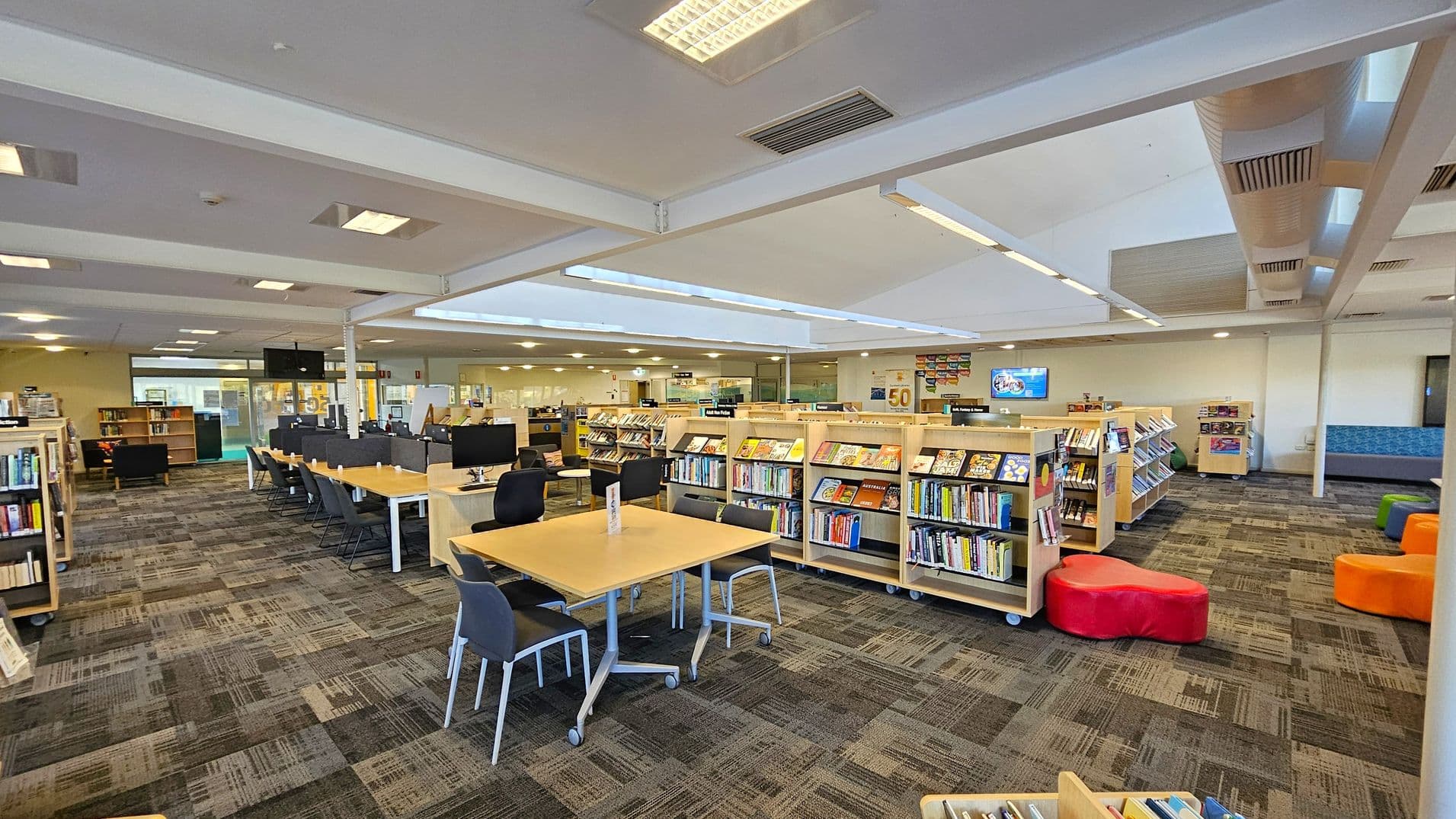 A spacious library interior with a patterned carpeted floor. On the left, there are rows of bookshelves filled with books. In the centre, a long table with computers and chairs is visible. To the right, more bookshelves are present, along with colourful seating poufs. The ceiling is fitted with fluorescent lights and air vents. The background features glass walls and additional seating areas.