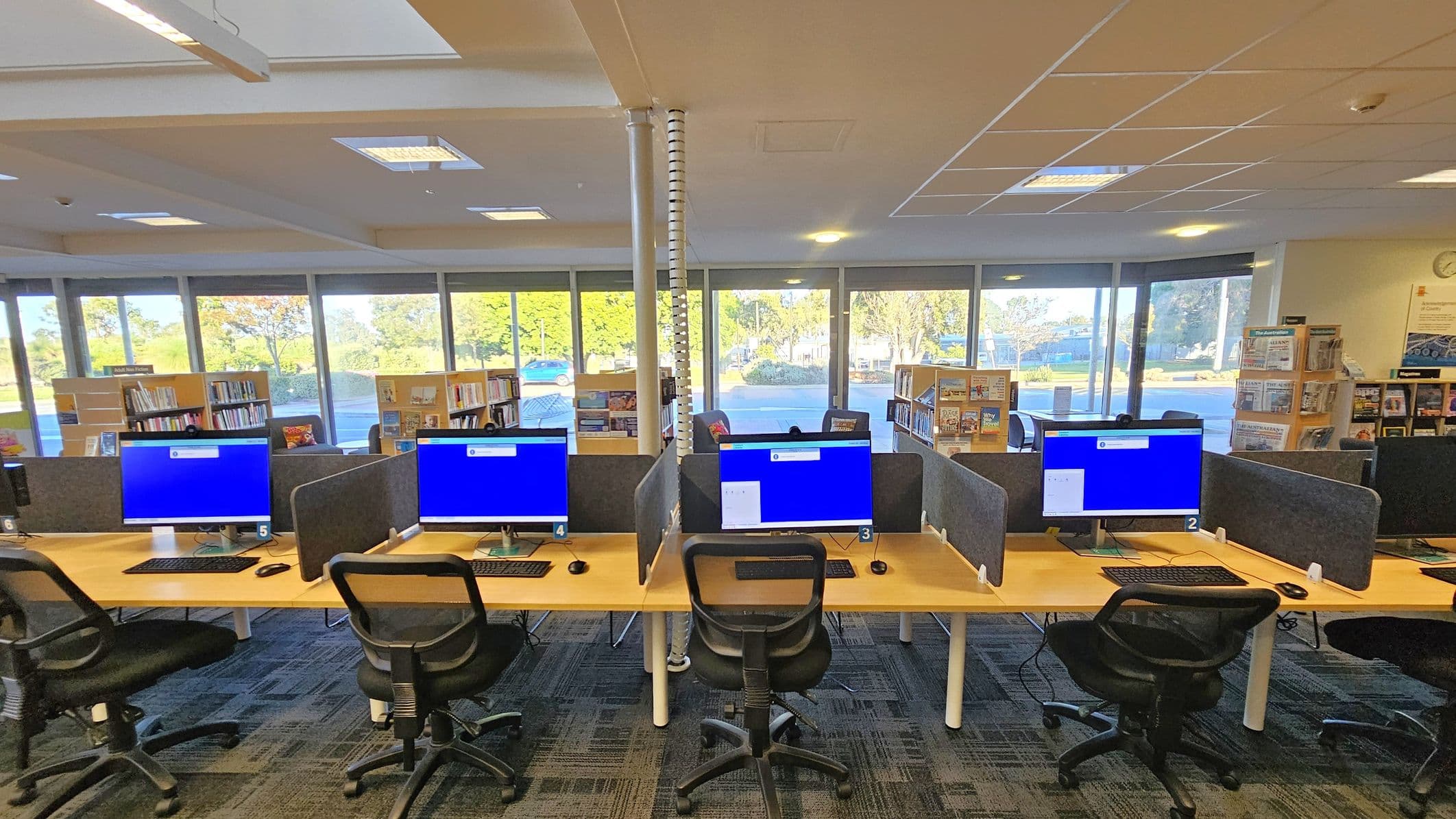 A library interior with a row of computer workstations in the foreground, each equipped with a monitor, keyboard, and mouse. Black office chairs are positioned at each station. The floor is carpeted with a patterned design. In the background, large windows allow natural light to illuminate the space, revealing bookshelves filled with books and magazines. The ceiling has a grid pattern with recessed lighting.