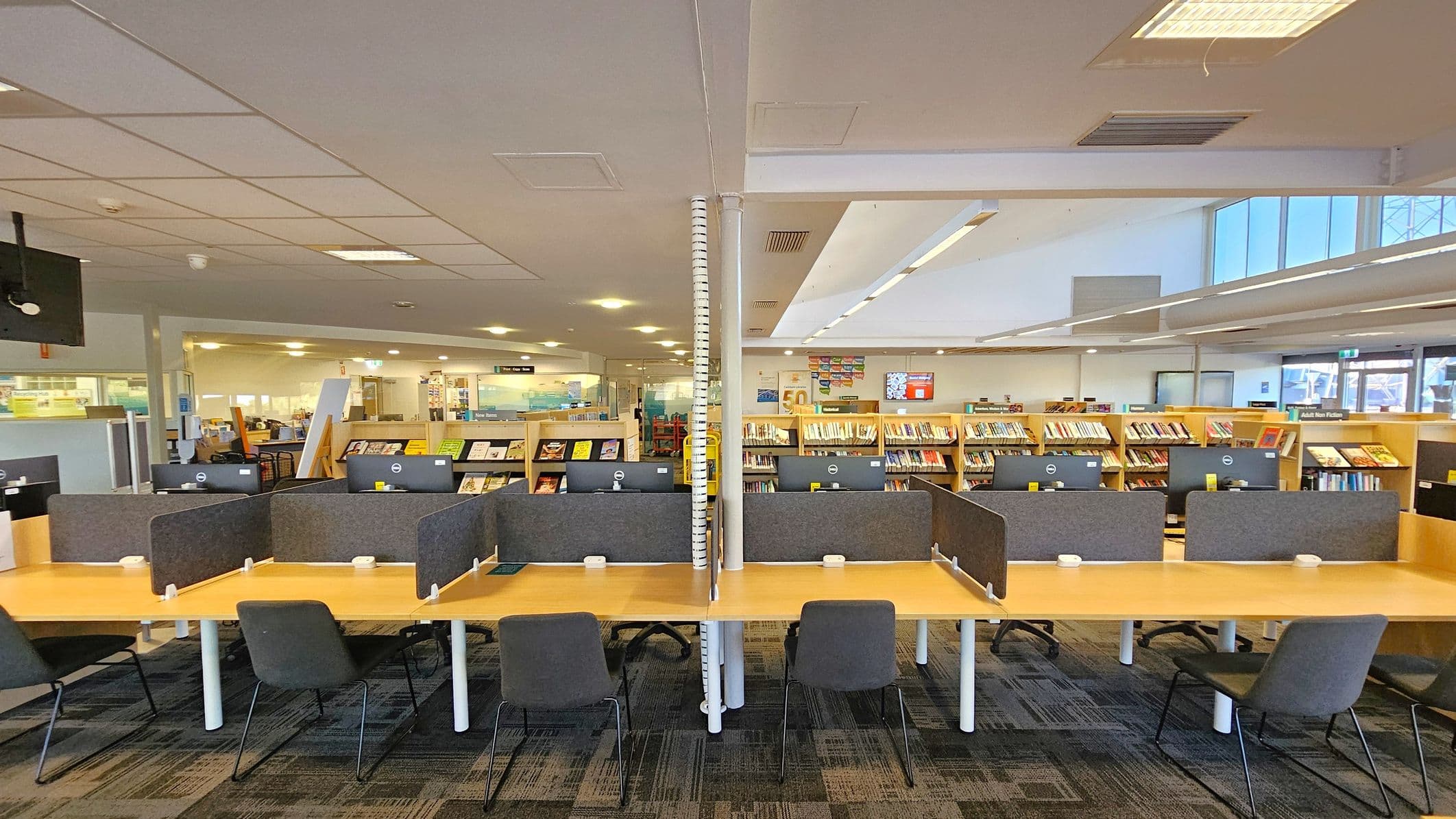 A library interior with a row of computer desks in the foreground, each equipped with a computer and surrounded by grey privacy dividers. Black chairs are placed at each desk. In the background, there are bookshelves filled with books. The ceiling is fitted with fluorescent lights, and the floor is carpeted with a patterned design. The space is well-lit and organised, with a modern and functional layout.