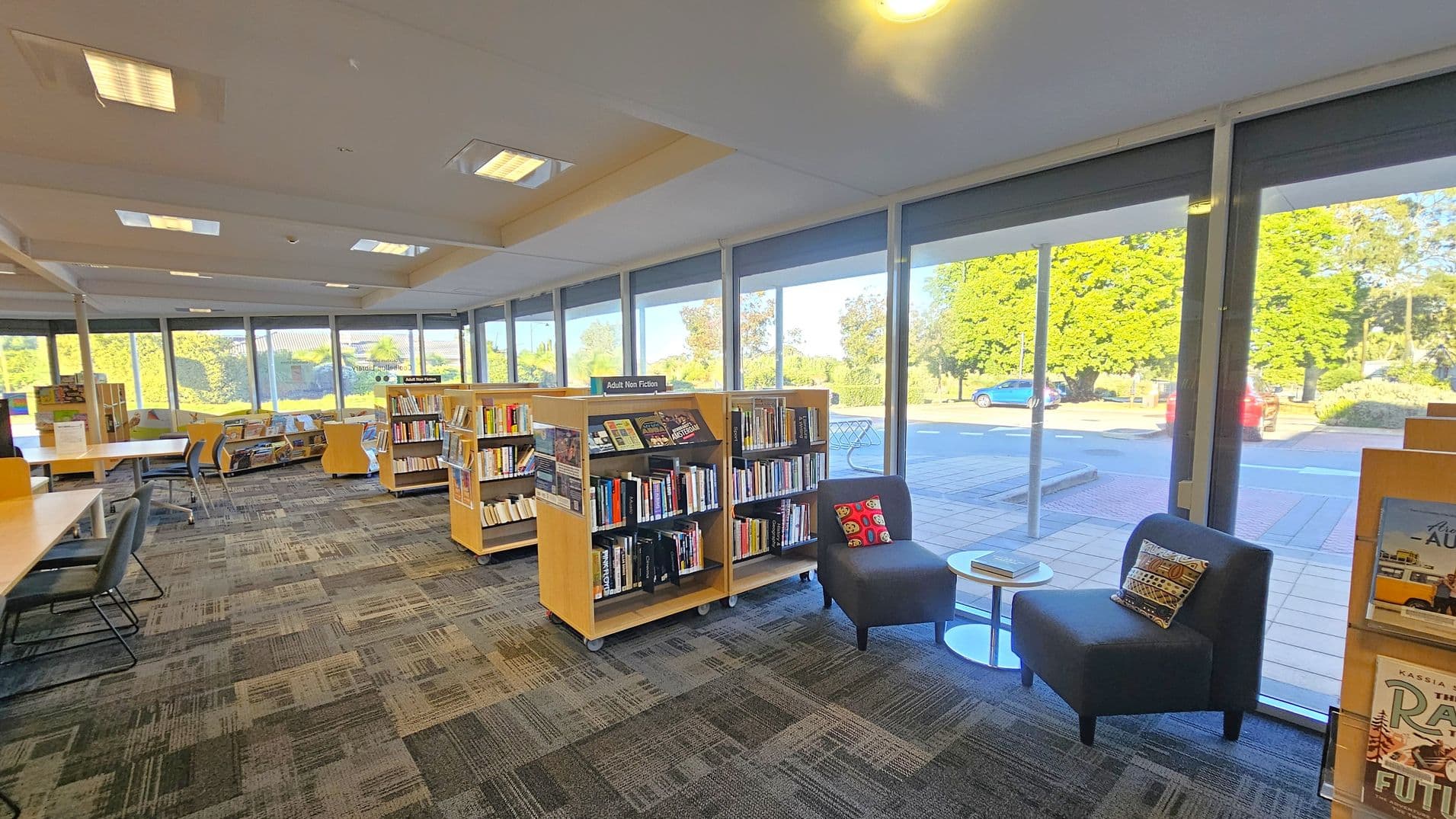 Inside a library with large floor-to-ceiling windows on the right, allowing natural light to illuminate the space. In the foreground, two grey chairs with patterned cushions are positioned around a small round table. Bookshelves labelled "Adult Non Fiction" are in the centre, filled with books. On the left, tables and chairs are arranged for reading or study. The floor is carpeted with a geometric pattern, and the ceiling has recessed lighting. Outside, trees and parked cars are visible.