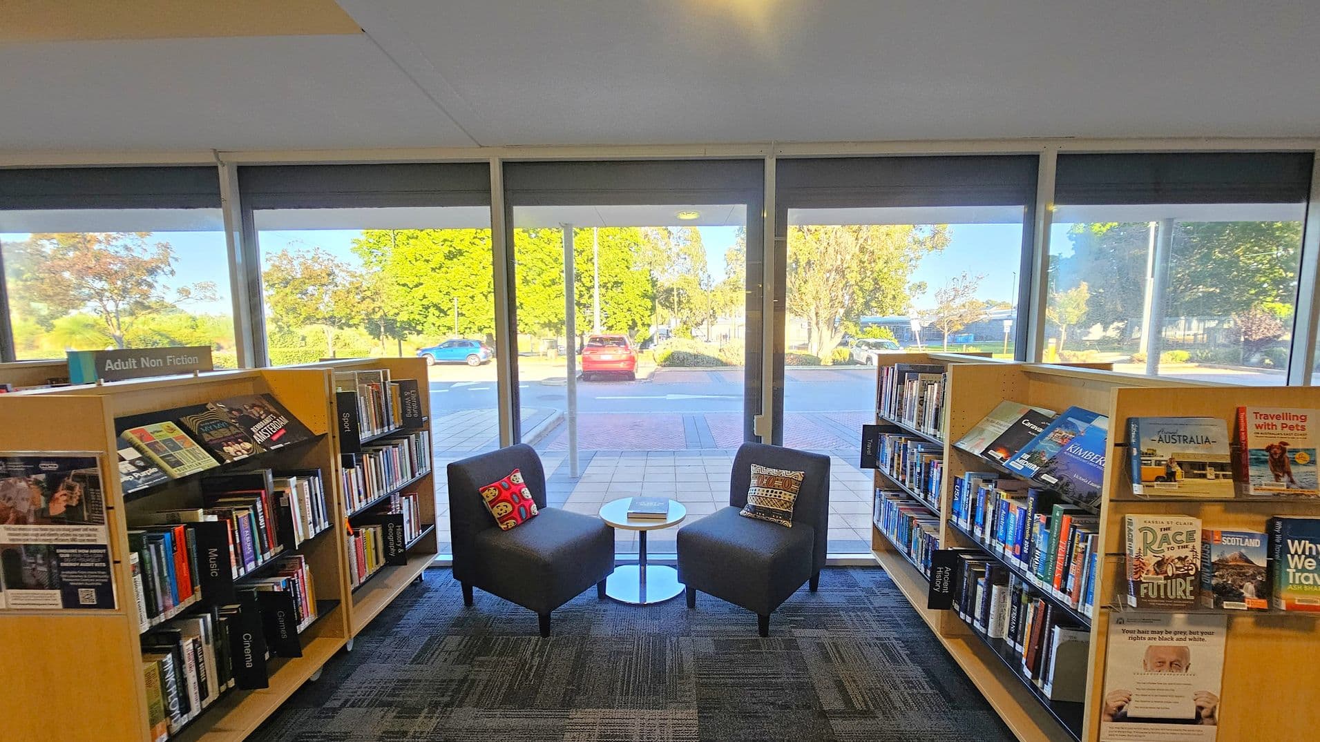 Bookshelves filled with books line the left and right sides, labelled "Adult Non Fiction." In the centre, two grey chairs with patterned cushions face each other, separated by a small round table with a book on it. Large windows in the background reveal a view of parked cars and greenery outside. The floor is carpeted with a geometric pattern, and the ceiling is plain with a light fixture.