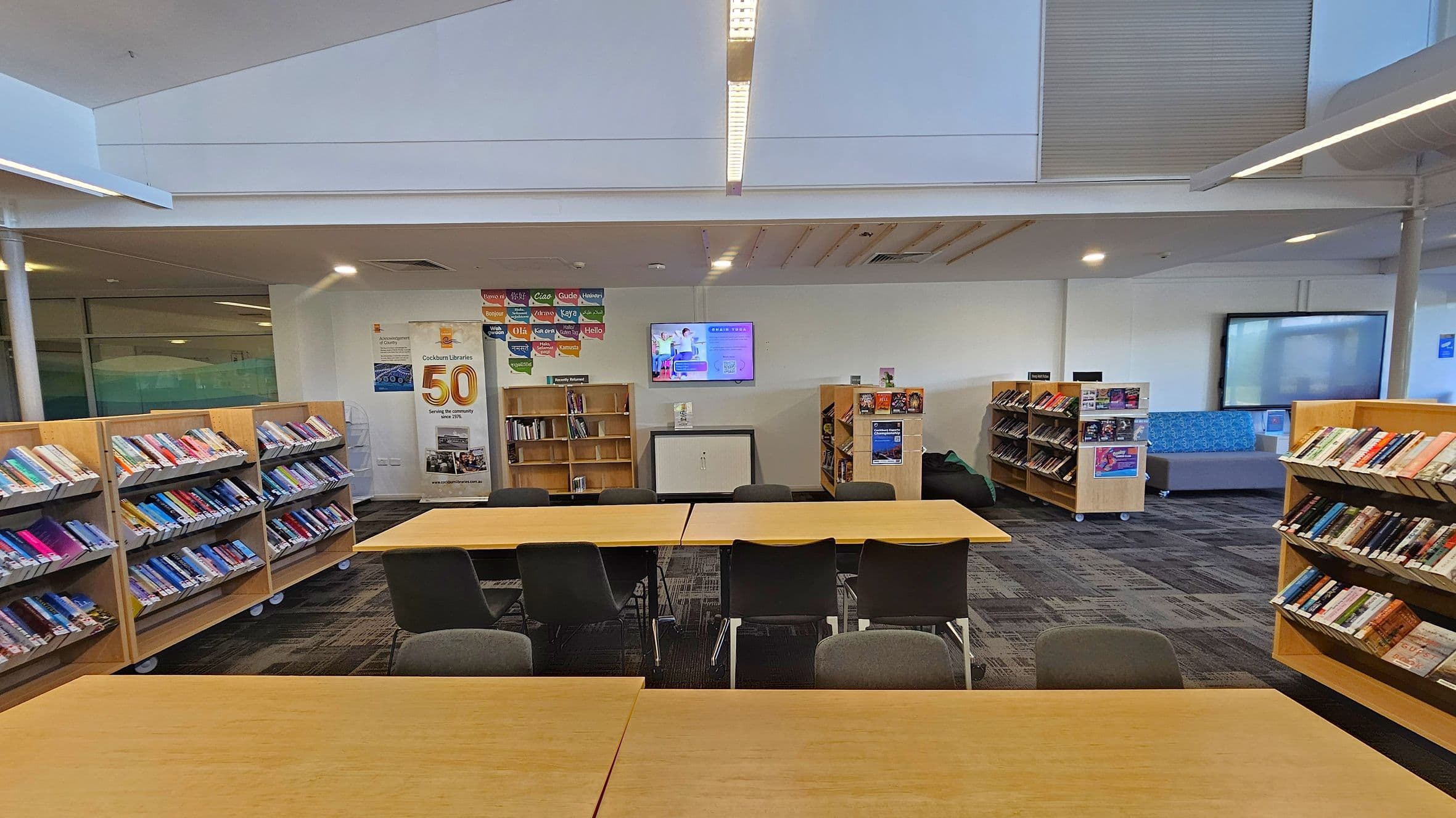 A library interior with wooden bookshelves filled with books on both the left and right sides. In the centre, there are tables with chairs arranged around them. A banner celebrating "50" years is displayed on the back wall, along with a television screen above it. The floor is carpeted in a dark pattern, and the ceiling has recessed lighting. A seating area with a blue patterned sofa is visible in the background on the right.