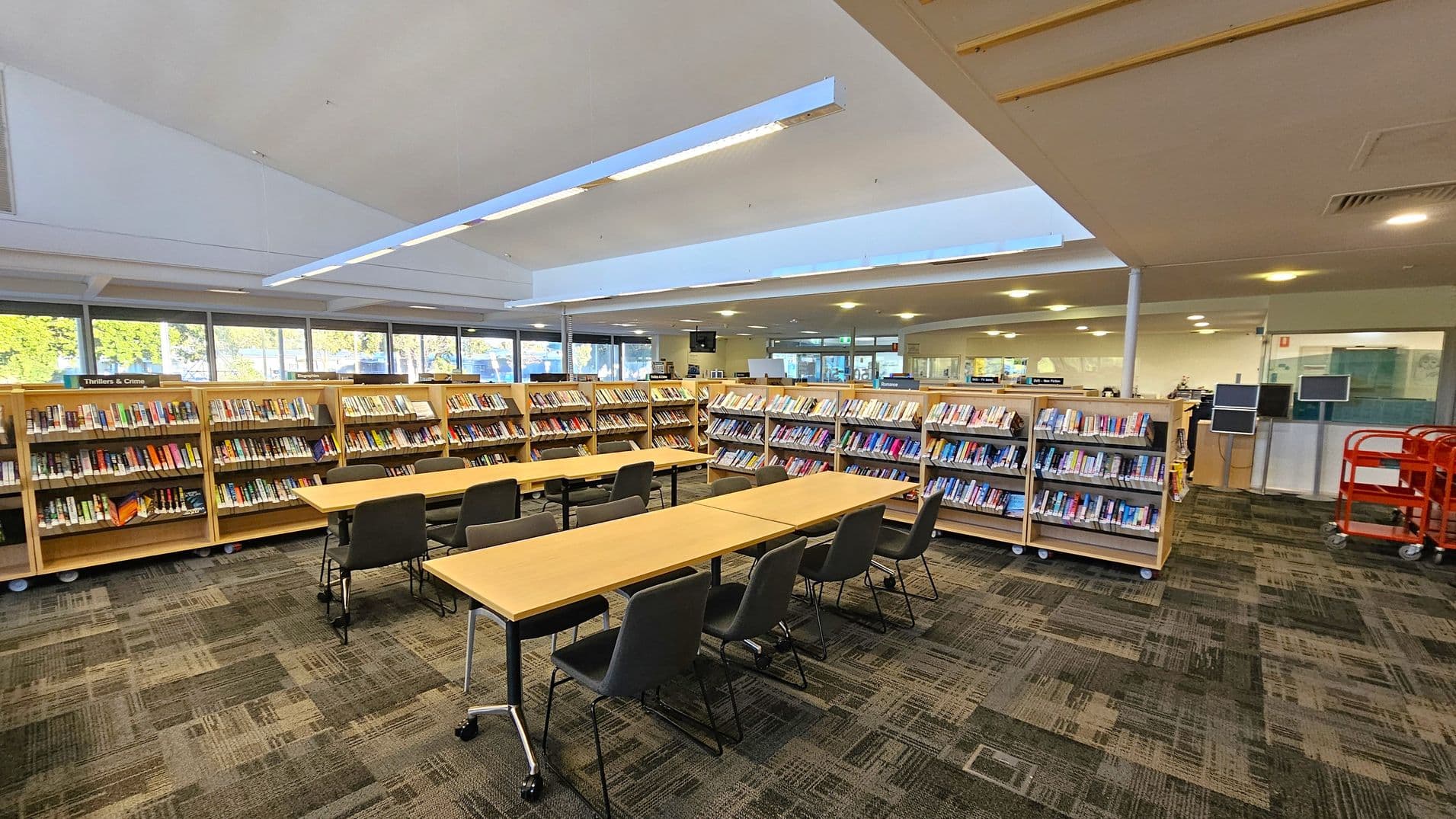 A library interior with bookshelves filled with books on the left and right. In the centre, there are long wooden tables with grey chairs arranged around them. The floor is covered with a patterned carpet in shades of grey and green. The ceiling is white with recessed lighting and a skylight. In the background, there are large windows allowing natural light in, and a few computer terminals are visible near the back wall. A red trolley is on the right side.