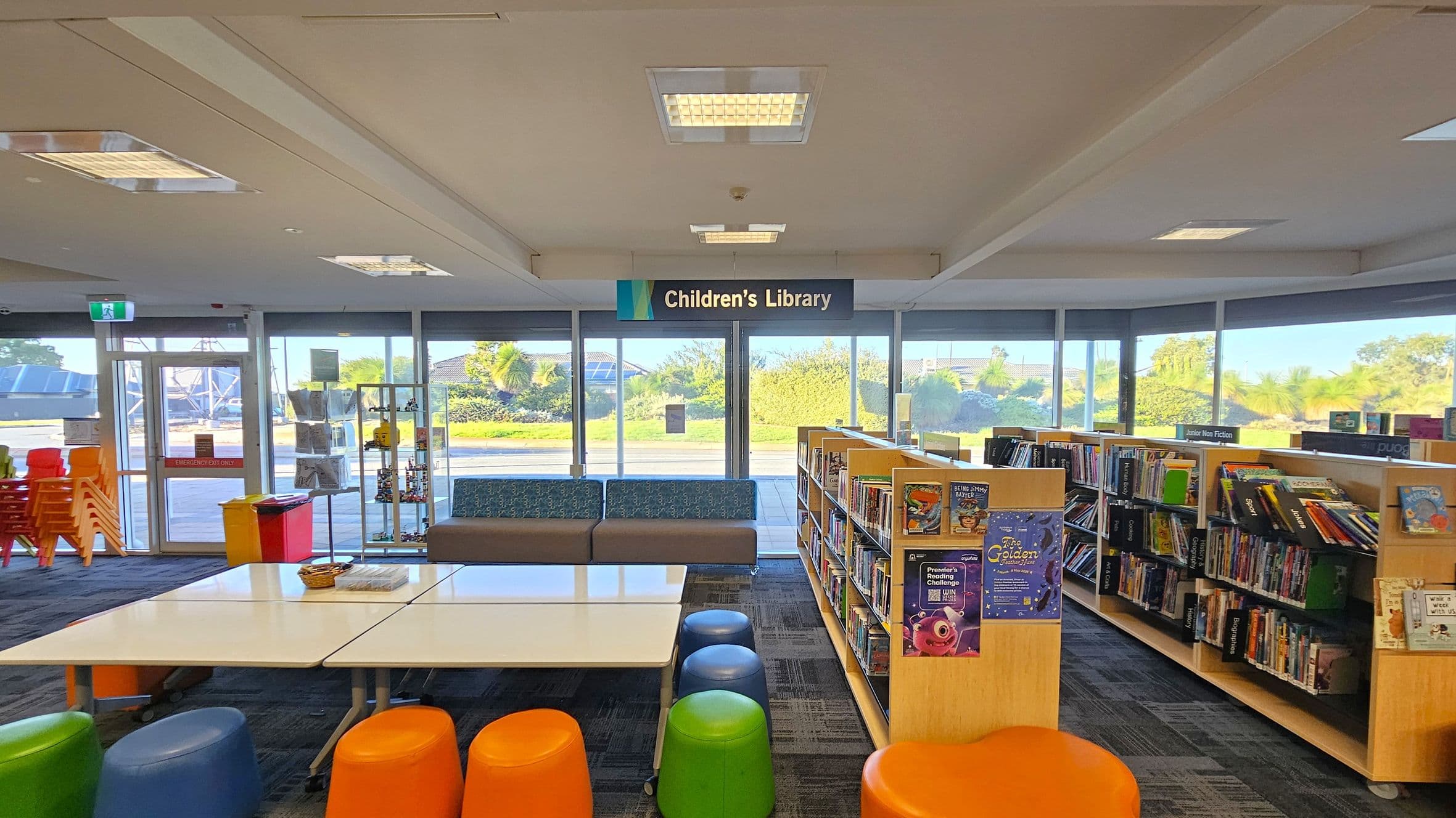 A children's library with a sign overhead. On the left, there are stacked colourful chairs and a yellow bin. In the centre, white tables are surrounded by colourful stools in orange, green, and blue. Behind the tables, a blue and grey sofa is positioned against large windows showing greenery outside. On the right, bookshelves are filled with children's books, labelled "Junior Non Fiction". The floor is carpeted with a patterned design, and the ceiling has recessed lighting.