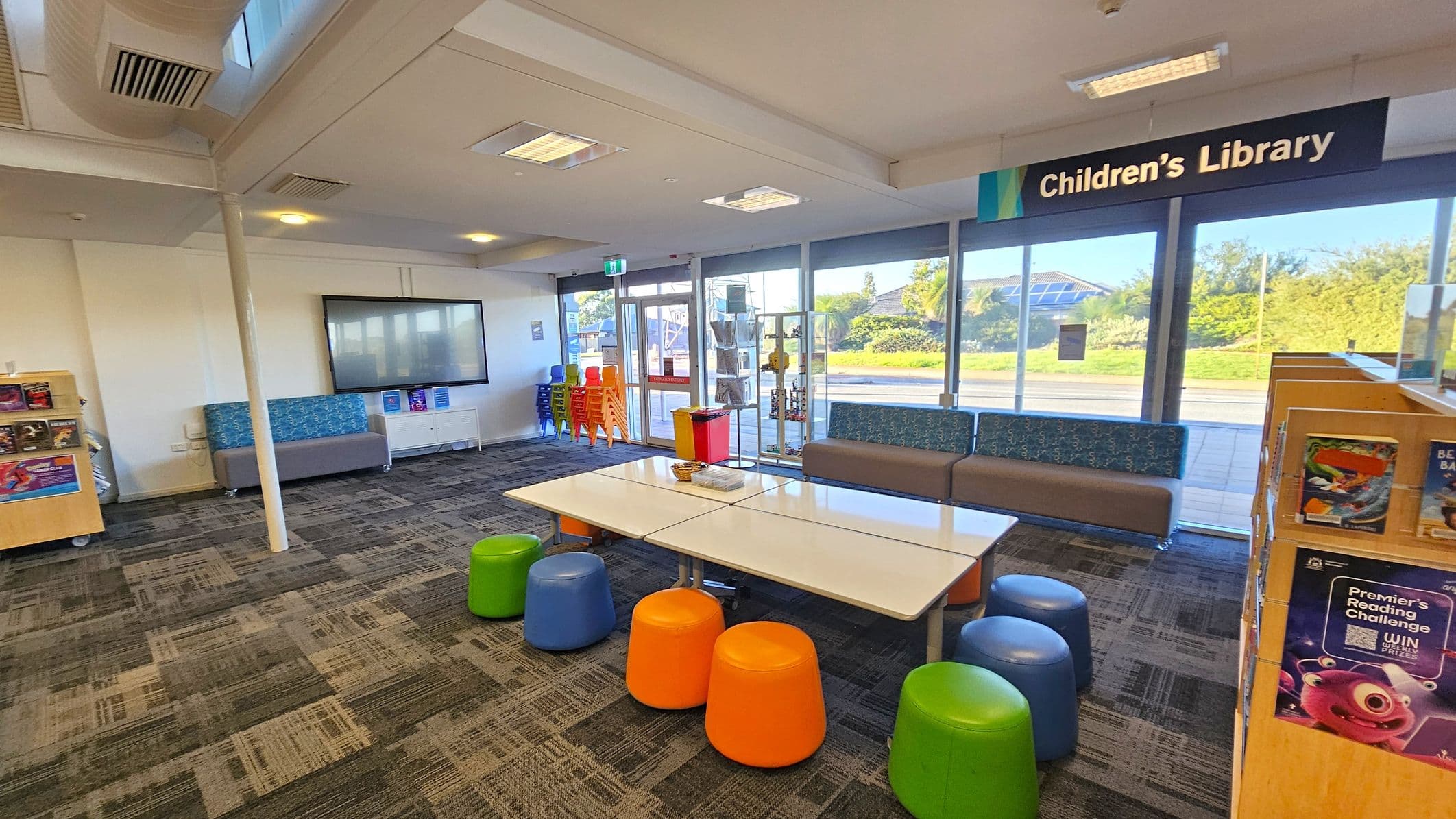 A children's library area with a sign overhead. On the right, there are bookshelves with children's books. In the centre, a table is surrounded by colourful stools in orange, green, and blue. The left side has a grey and blue sofa against the wall, with a large screen above it. The floor is carpeted with a patterned design, and the ceiling has recessed lighting. Large windows in the background let in natural light, showing greenery outside.