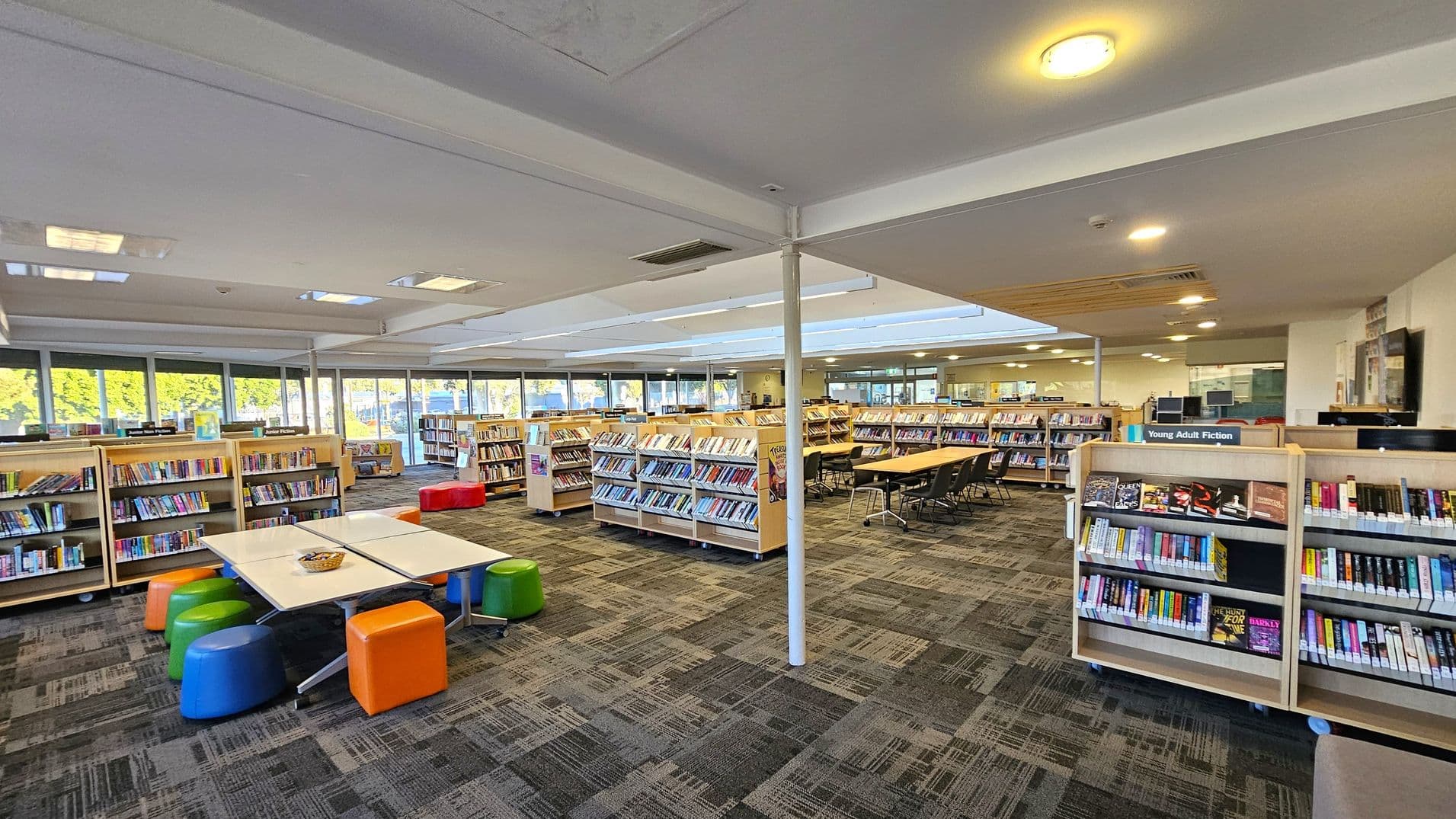 A spacious library interior with bookshelves filled with books on both sides. In the foreground, there is a table with colourful stools in blue, green, and orange. The floor is carpeted with a patterned design. The ceiling is white with recessed lighting. Large windows in the background allow natural light to enter, and there are more bookshelves and tables with chairs in the middle and background areas. A sign on one shelf reads "Young Adult Fiction."