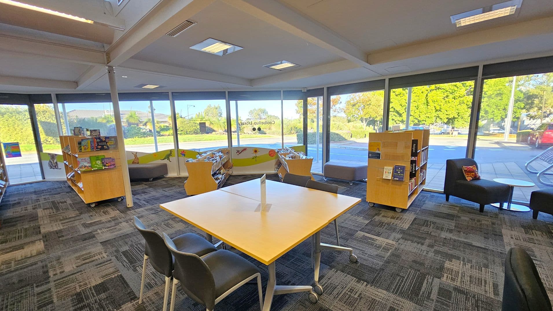 A library interior with large glass windows in the background, offering a view of greenery and a parked red car outside. In the foreground, a square table with four chairs is positioned on a patterned carpet. To the left and right, there are wooden bookshelves filled with books. A black armchair with a cushion and a small round table are on the right. The ceiling features recessed lighting.