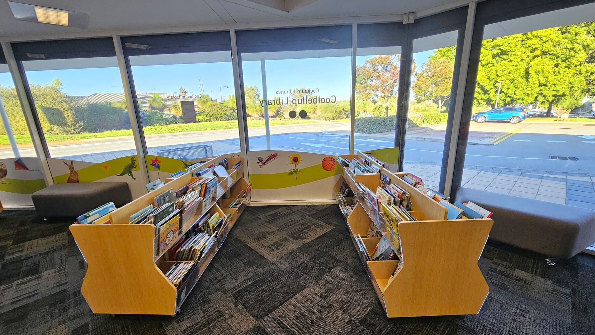 Inside a library, two wooden bookshelves filled with children's books are positioned in a V-shape. The floor is carpeted with a geometric pattern. Large windows in the background provide a view of the outside, showing a road, greenery, and a blue car. The windows have colourful decals featuring a kangaroo, dinosaur, aeroplane, sunflower, and basketball. Grey cushioned seating is visible along the windows.